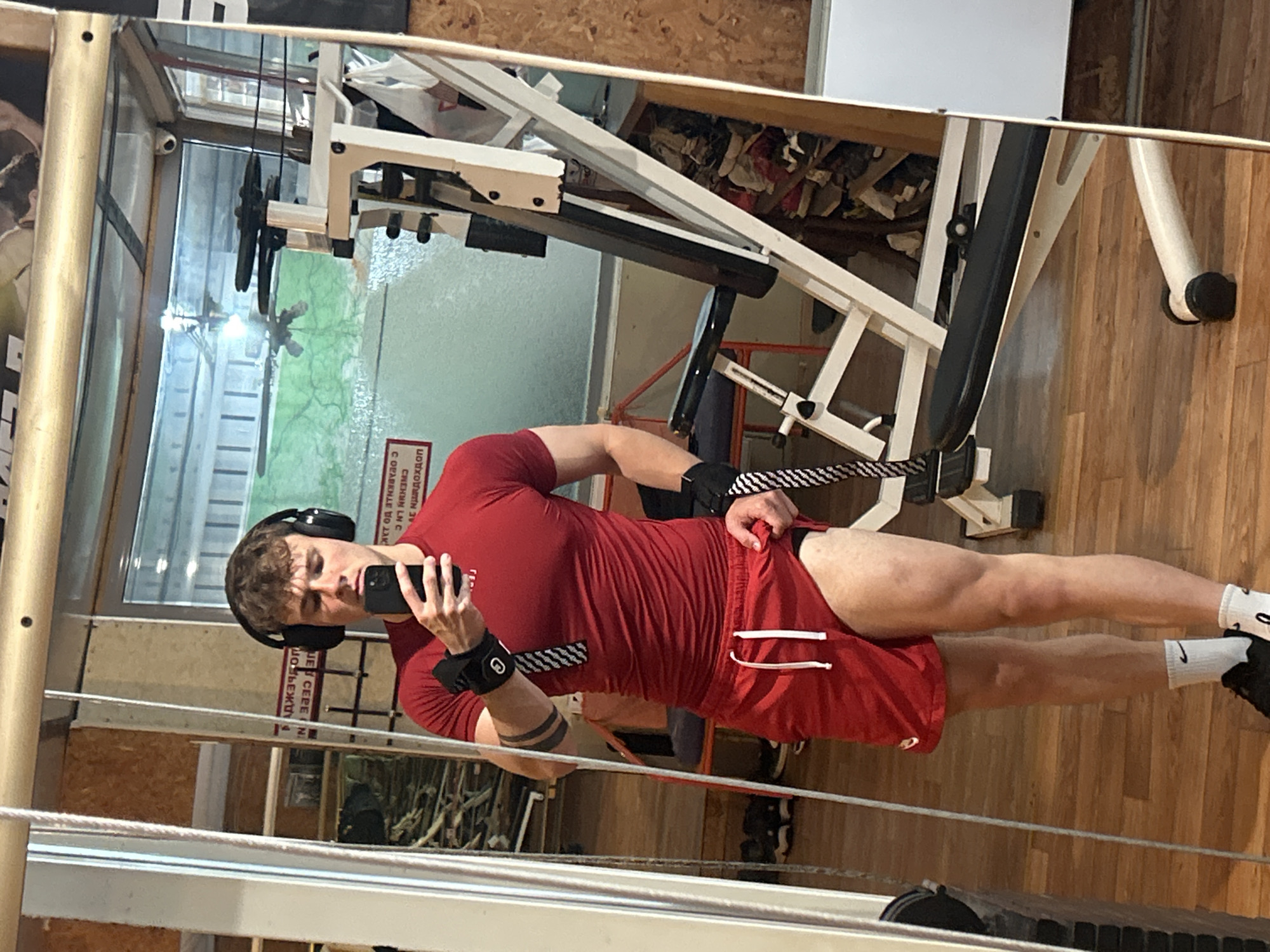 A man wearing red workout clothes, headphones, and athletic wristbands is taking a selfie in a gym. He is positioned in front of a mirrored wall, likely inside a fitness center in Elhovo, Bulgaria. The gym appears to be equipped with standard weightlifting machines and equipment. The background includes a view of the outdoors, possibly a park or residential area, seen through a window. The scene is well-lit, suggesting daytime or effective artificial lighting.