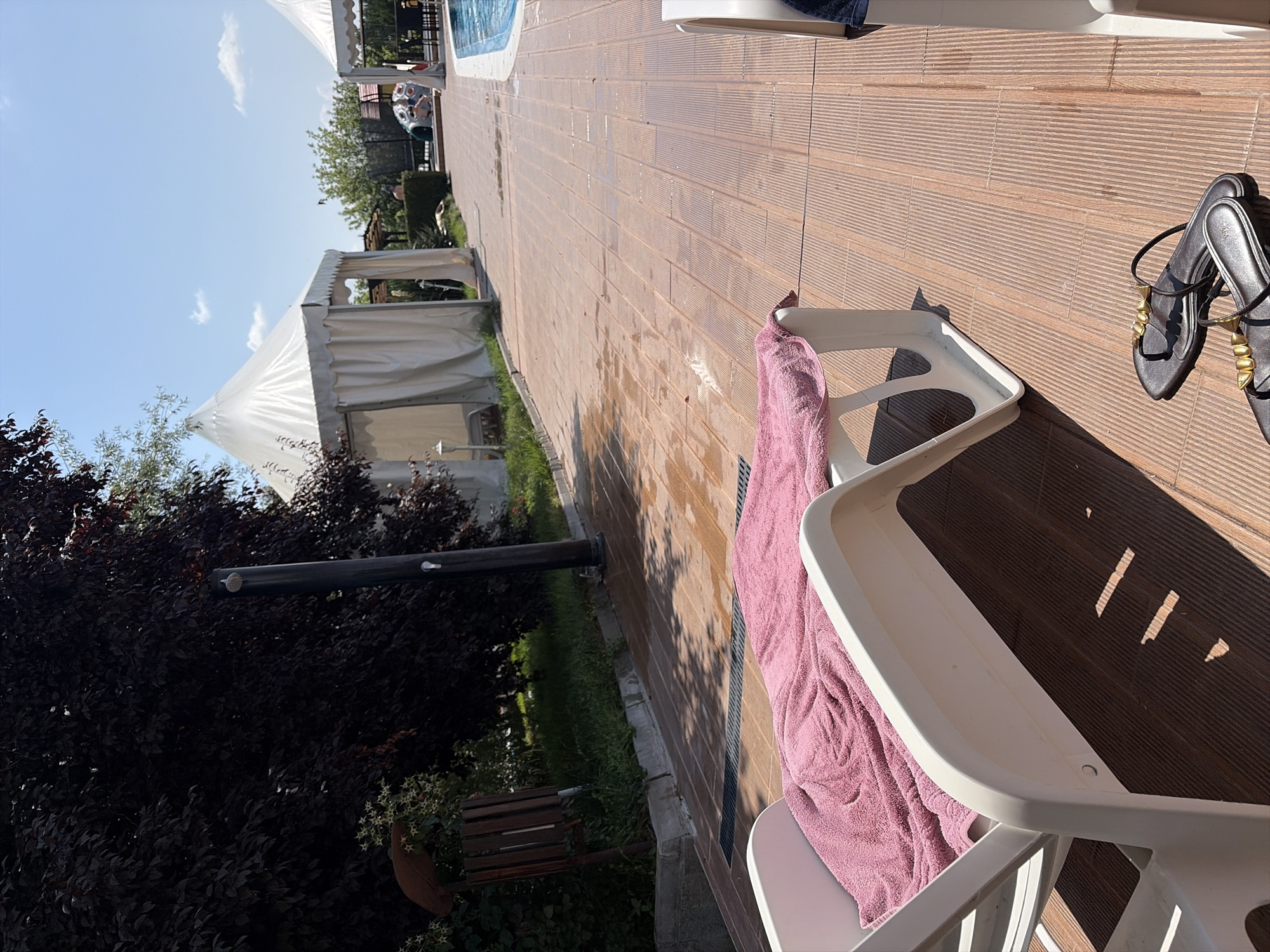 The image depicts an outdoor scene, likely a resort or recreational area, in Simeonovo, Bulgaria. In the foreground, a white chaise lounge is covered with a pink towel. To the right of the chaise lounge, a pair of dark-colored sandals with gold accents lies on the wooden deck. The deck appears to be wet in places, suggesting recent activity like swimming or cleaning.
In the midground, a dark-leaved tree dominates the left side of the frame, with a black shower pole positioned near its trunk. Beyond the tree, a white, tent-like structure with open sides is visible. Further back, another similar tent is present, and a swimming pool is partially visible on the right. Several people are sparsely scattered in the background, with some near the pool and others near the tent structures. The sky is clear and blue with a few scattered clouds, indicating a sunny day. The overall environment suggests a leisure setting.