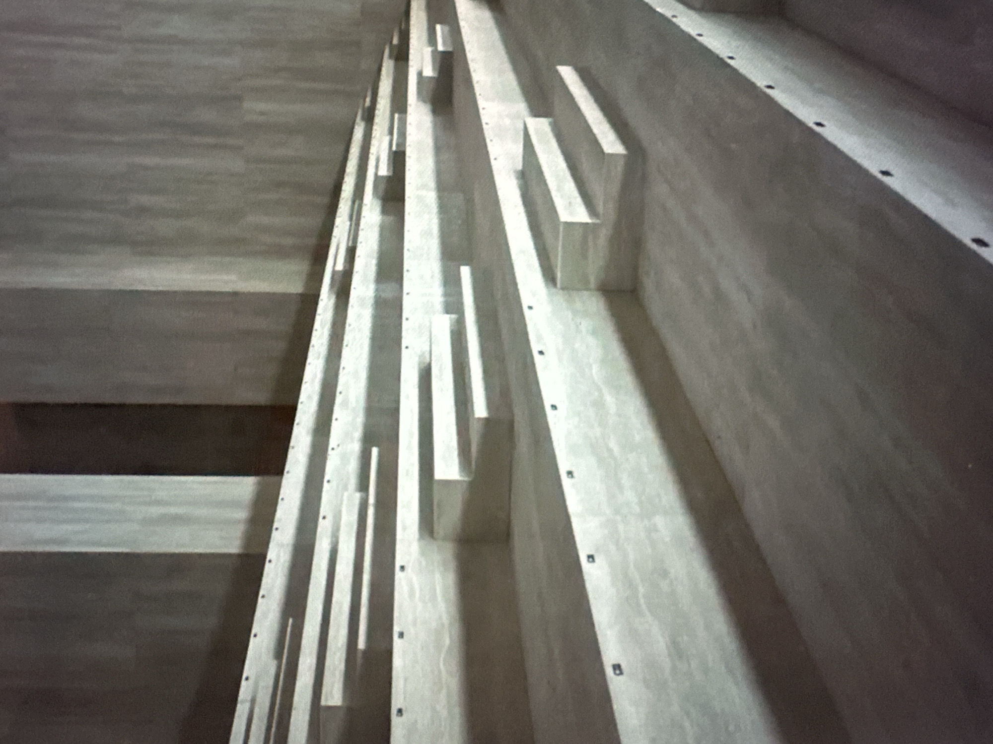 The scene depicts an empty, minimalist interior architectural space located in the city of Wealden, United Kingdom. The primary features are a series of ascending, parallel horizontal tiers constructed from a light-colored, textured material, possibly concrete or stone with a veined pattern. Six prominent tiers are visible, receding from the lower right foreground towards the upper left background. Small, dark, rectangular fixtures are embedded at regular intervals along the front edge of each tier. At various points on these tiers, smaller rectangular blocks form individual steps, creating multi-level seating or access points. These blocks cast distinct shadows, indicating illumination from an unseen overhead or side source. The background consists of tall, vertical wall sections or pillars made of the same material, exhibiting subtle variations in light and shadow. The environment is devoid of people, objects, actions, or interactions.