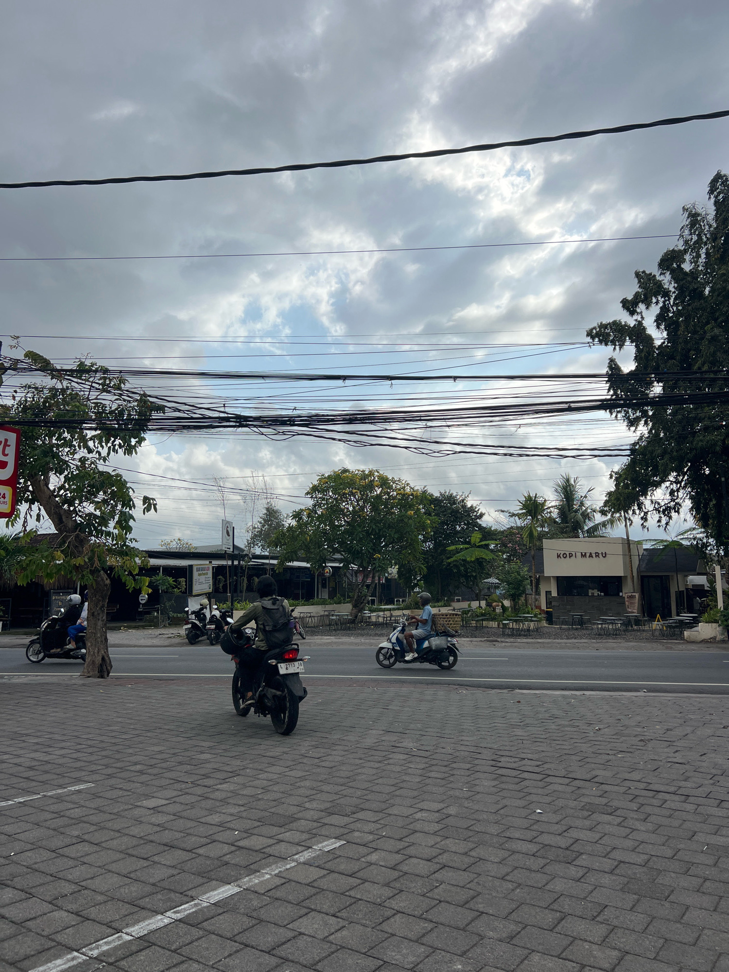 The image depicts a street scene in Denpasar, Indonesia, likely in a commercial area. The foreground shows a gray brick-paved walkway with white painted lines, suggesting a pedestrian area or parking spaces. Several motorcycles are visible on the road in the middle ground. One motorcycle in the center, facing away from the camera, is ridden by a person wearing a green backpack. Another motorcycle to the left is carrying two people. Further to the right, a single rider on a white motorcycle is heading towards the left.
Along the roadside, there are various establishments, including a building with signage that reads "KOPI MARU." There are outdoor seating areas with tables and chairs in front of some of these establishments. Lush green trees and vegetation line the street, contributing to a tropical atmosphere. Overhead, a dense network of electrical wires crisscrosses the sky, supported by poles that are not fully visible. The sky is overcast with scattered clouds, indicating a possibly cloudy or approaching rainy day. The overall impression is that of a busy street with ongoing daily activities in Denpasar.