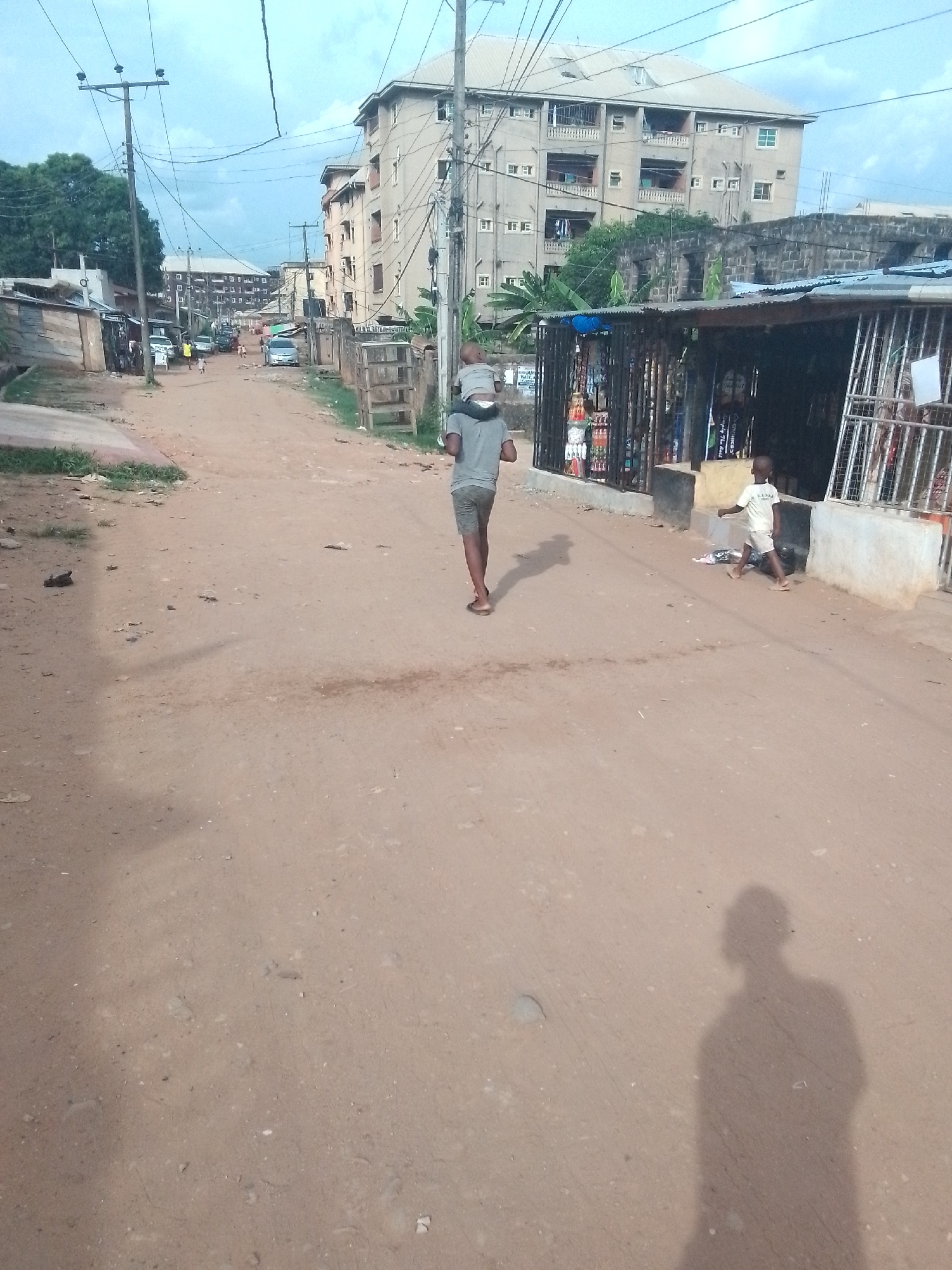 The image depicts a dirt road in an urban environment. A man is carrying a child on his shoulders, walking towards the viewer. Another child is running near a small shop on the right side of the road. Buildings are visible in the background, and power lines run overhead. The environment is dusty and appears to be a residential area.