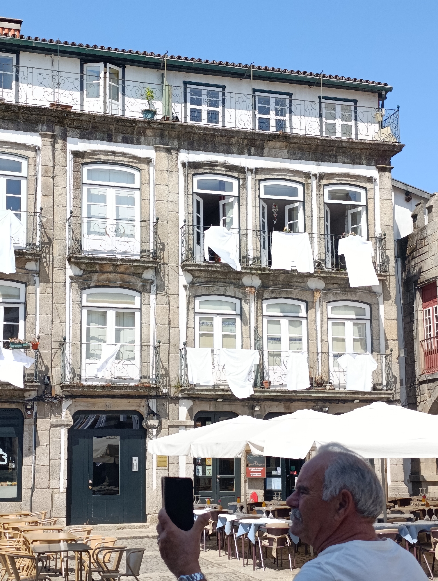 The image captures a street view in Guimarães, Portugal, featuring a weathered stone building with multiple floors and balconies. White laundry is hanging to dry from the balconies, contrasting with the building's facade. On the ground floor, outdoor cafe tables and chairs are set up, suggesting a public space. In the foreground, a man is seen from the side, holding up a smartphone to take a picture of the building. The man has gray hair and a mustache, and is wearing a white t-shirt. The sky above is clear and blue. The architecture of the building, with its stone construction, arched windows, and decorative iron railings on the balconies, is indicative of historical European urban environments. The presence of cafe seating and the man taking a photo suggests a tourist or local enjoying the outdoor ambiance.