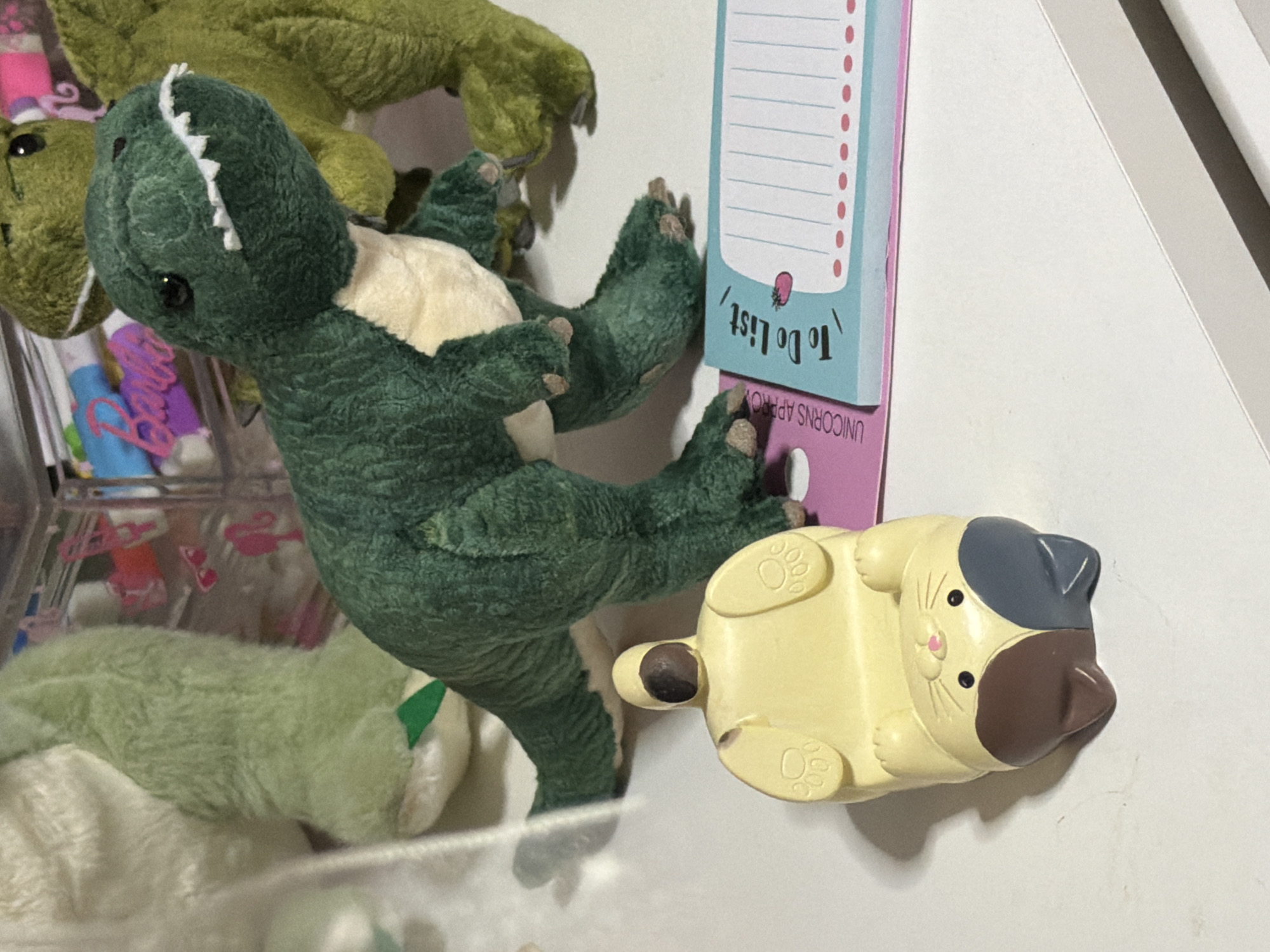 A close-up shot shows a plush toy dinosaur in dark green with a cream-colored belly and sharp white teeth, positioned on a white surface. Behind it, another, lighter green plush toy dinosaur is partially visible. To the left of the main dinosaur, a yellow cat-shaped object with paws and a brown and gray face is lying on its back, with its legs pointing upwards. In the foreground, a blue and pink notepad with the words "To Do List" and a drawing of a strawberry is laid out. The background shows a blurry pink and blue object with the word "Barbie" in pink script, along with other colorful items in a clear container. The scene appears to be indoors, on a flat surface, possibly a desk or shelf. There is no indication of the city of Moscow, Russia, in the image.