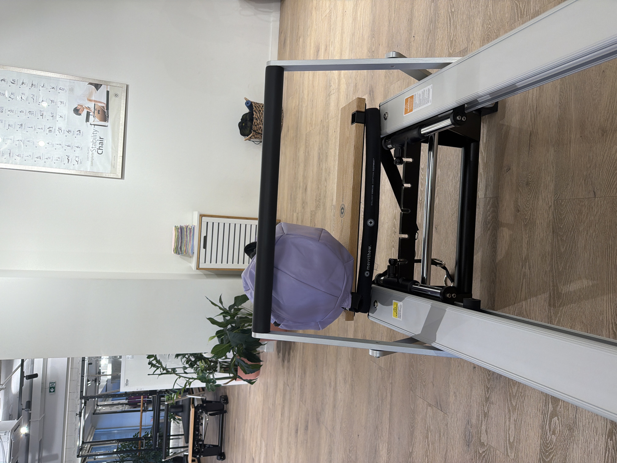 In the city of Boreti, Montenegro, a well-lit indoor fitness studio features a pilates reformer machine in the foreground. The machine is silver and black, with a wooden platform on top. A light purple yoga ball is resting on the platform. In the background, there is a white wall with a framed poster depicting pilates exercises. To the left of the wall, a white cabinet with drawers is visible, with a stack of colorful towels on top. Further in the background, a plant with large green leaves sits in a pink pot. Another pilates apparatus is visible in the far left background. The floor is made of light wood planks. A woven basket filled with dark items is placed on the floor near the wall. The overall impression is of a clean, organized, and professional fitness space.