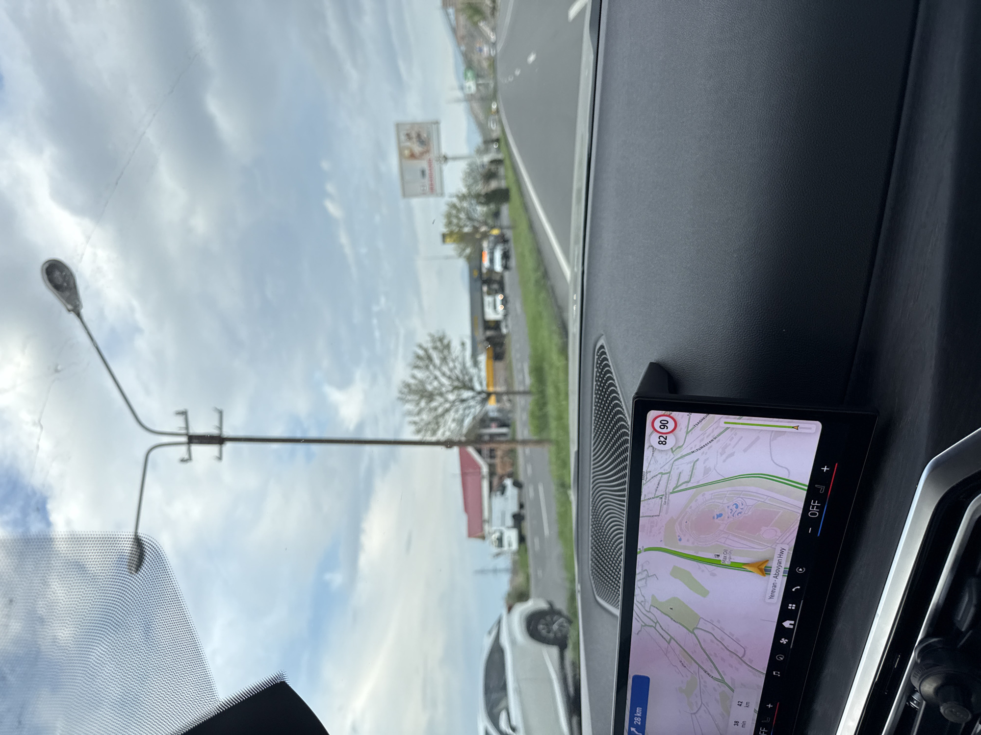 The view from inside a car shows a road leading towards Yerevan, Armenia, with a GPS navigation display indicating the driver is on the Yerevan-Abovyan Highway and approximately 28 km from the city center. The surrounding environment includes roadside infrastructure such as streetlights and billboards. A white car is visible in the foreground. The sky is cloudy. The GPS display shows a speed limit of 90 and the current speed as 82.