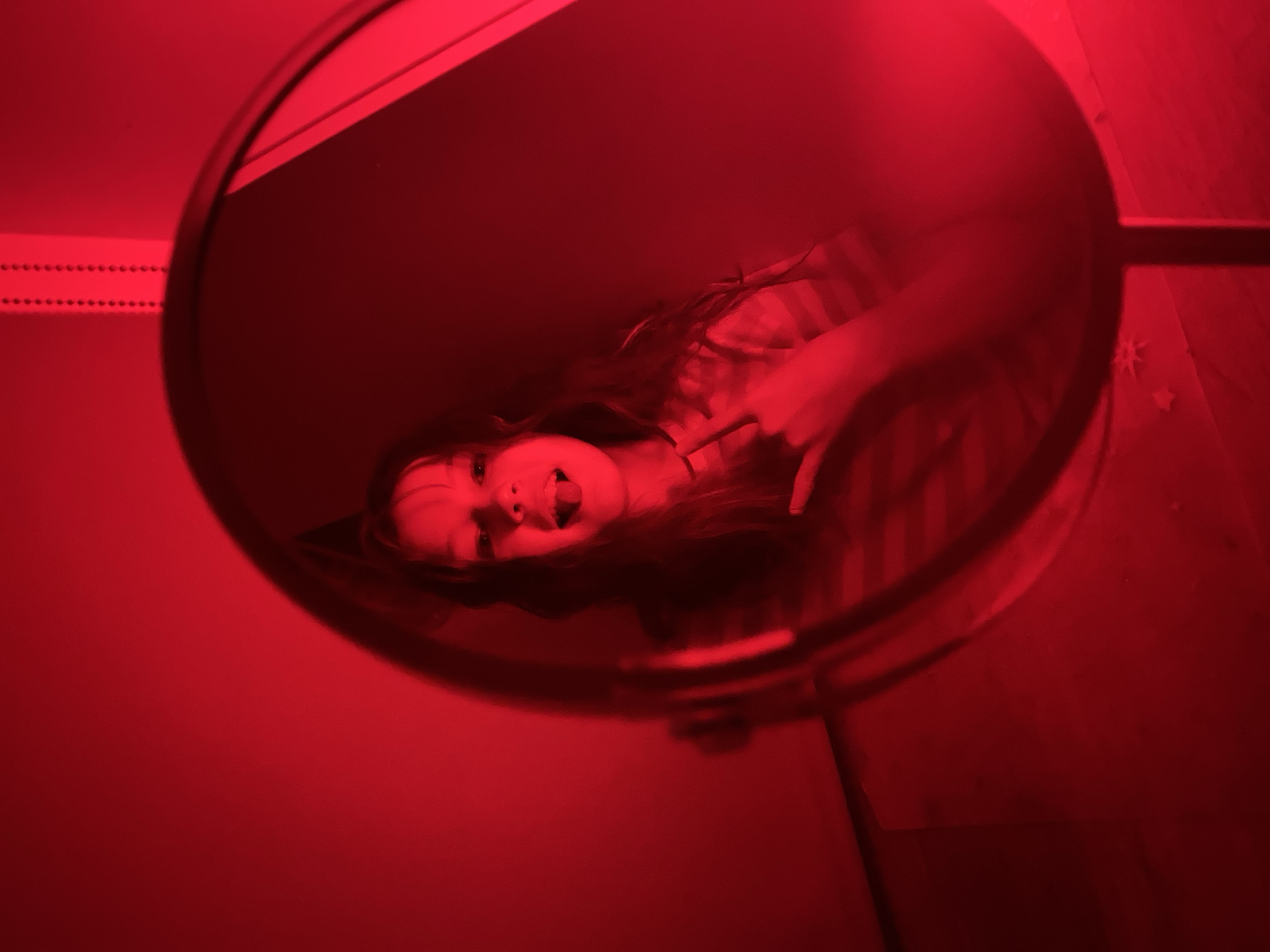 A young person is visible in the reflection of a round mirror. The person is sticking their tongue out and pointing a finger. The scene is bathed in red light, suggesting a red light source. The mirror is positioned against a red wall. A strip of dotted lights is visible on the upper left of the mirror. The bottom right of the mirror reveals a portion of a wall and a few star-shaped decorations. The location is Chaikovsky, Russia.