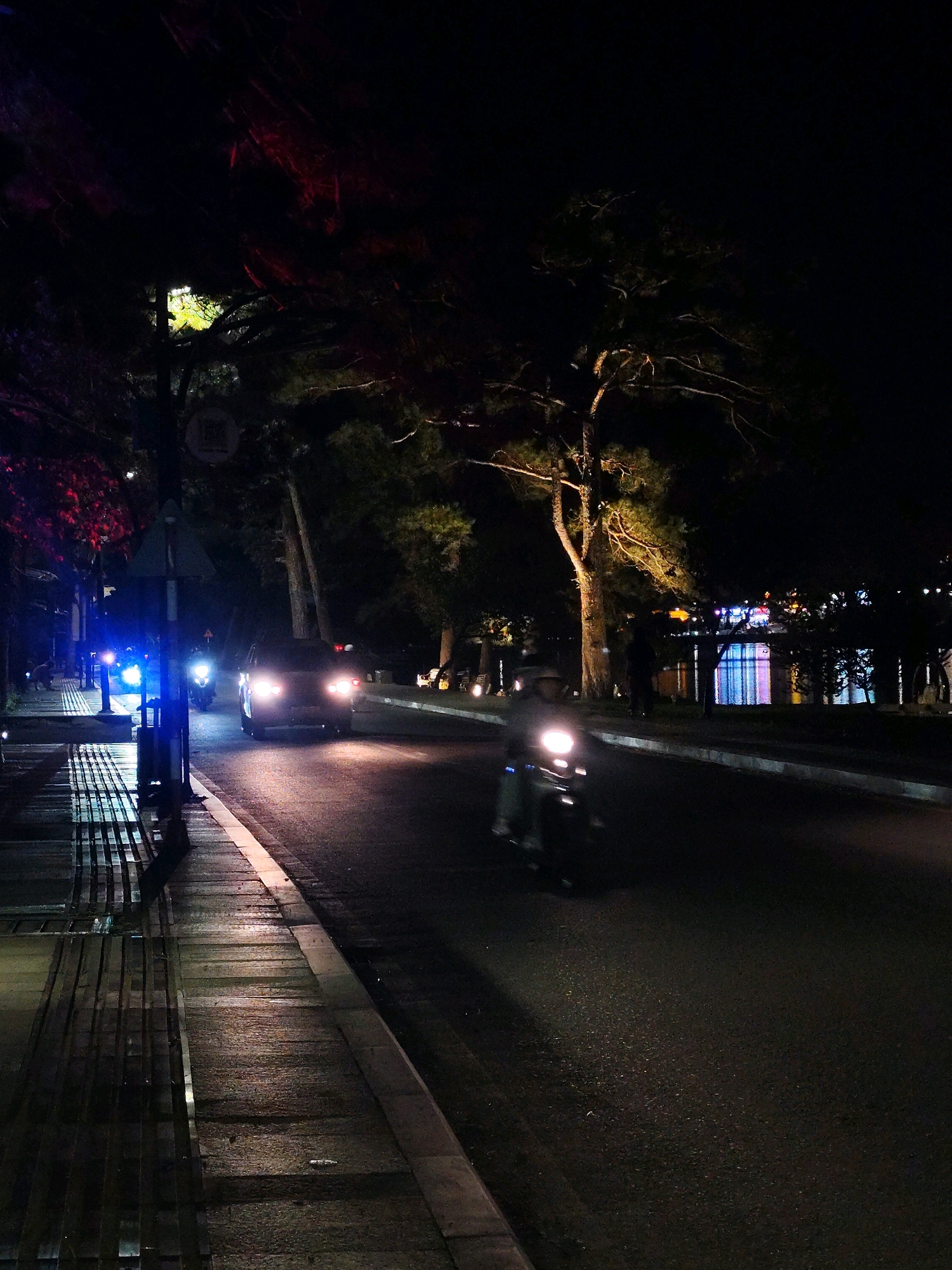 The image depicts a street scene at night in Xuân Hương - Đà Lạt, Vietnam. A paved road runs horizontally across the frame, with a sidewalk on the left. Several vehicles are present: a motorcycle with its headlight illuminated is in the foreground on the right side of the road, and a car with its taillights on is in the middle ground on the left side of the road. Another motorcycle, also with its headlight on, is further down the road. The sidewalk appears wet, reflecting the lights from street lamps and the vehicles. Trees line the street, with some foliage illuminated by artificial lights, casting shadows. In the background, buildings with colorful illuminated windows are visible across a body of water, suggesting a lakeside or riverside location. The overall atmosphere is dark, with artificial lights creating pools of illumination on the wet surfaces.