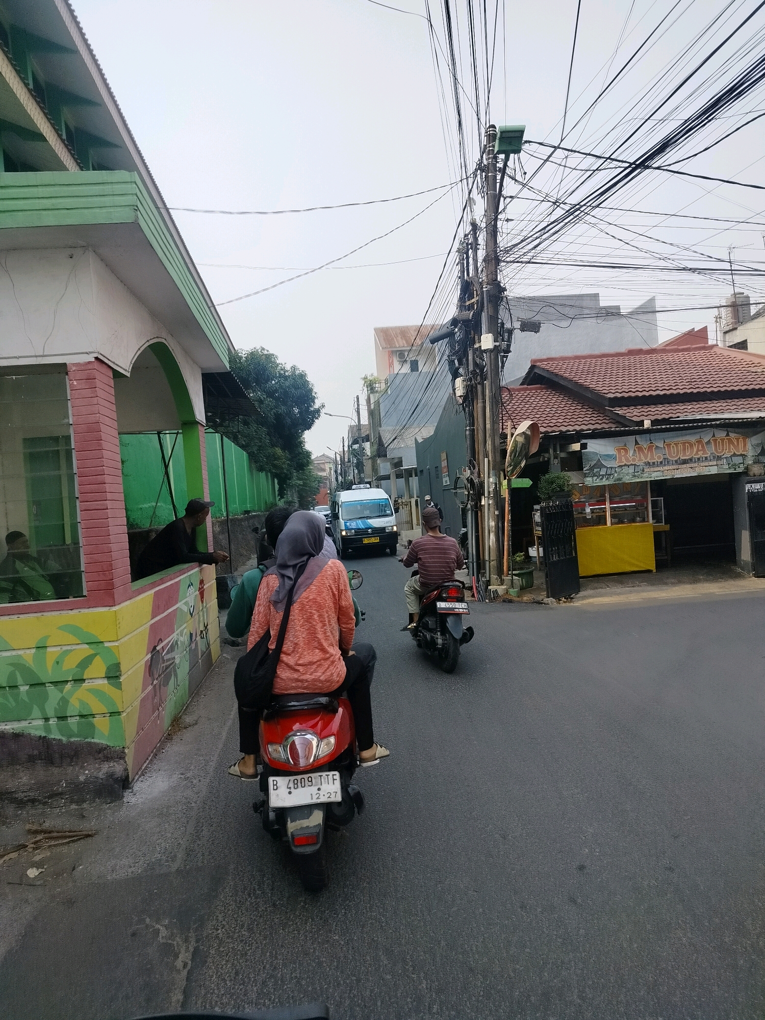 In East Jakarta, Indonesia, a street scene unfolds under an overcast sky. The foreground features the rear of a red scooter with two occupants: a woman wearing an orange shirt and a grey hijab, and another person behind her wearing a black shirt. The scooter's license plate reads "B 4809 TTF 12-27". To the left, a building with a green and white facade features colorful murals along its base. Several men are visible within the covered area of this building, with one leaning out.
The street is paved and appears to be wet in some areas. Further down the road, a white van is visible, and to its right, a street pole is heavily laden with tangled electrical wires, characteristic of urban Indonesian infrastructure. Adjacent to the pole is a building with a brown tiled roof, housing a food stall labeled "R.M. UDA UNI". The background shows more buildings and trees. The overall impression is a typical day on a busy street in an Indonesian city.