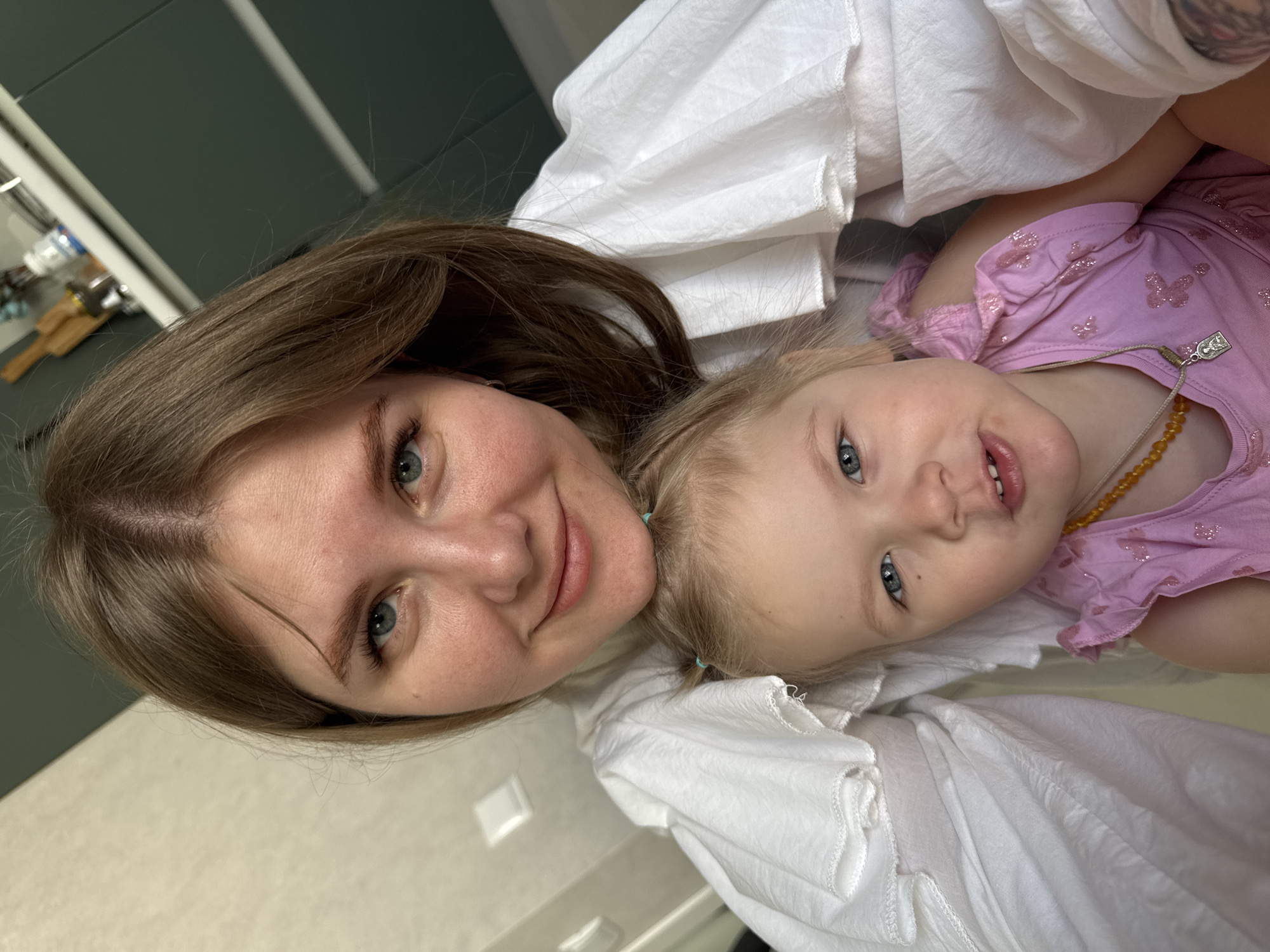 This image captures an adult female and a young female child positioned closely together, likely in a selfie, within a residential interior in the city of Gornyi (Dvurechenskoe S/P), Russia. The adult, positioned behind and above the child, has light brown hair parted centrally, blue eyes, and is looking towards the camera with a slight smile. She is wearing a white blouse with visible ruffles on the shoulders. The child, a toddler, is in the foreground, with blonde hair styled into two small pigtails secured by light blue hair ties. She has blue eyes, a small dark mark below her right eye, and her mouth is slightly ajar. She is wearing a pink sleeveless top with subtle sparkle patterns and a yellow bead necklace with a small silver-toned pendant. The background on the left consists of a light-colored wall with a white electrical outlet or switch plate. The background on the right, behind the adult's head, shows dark green cabinetry, with a partial view of a shelf above it displaying a wooden rolling pin, a white bottle, and other household items, indicative of a kitchen or utility area.