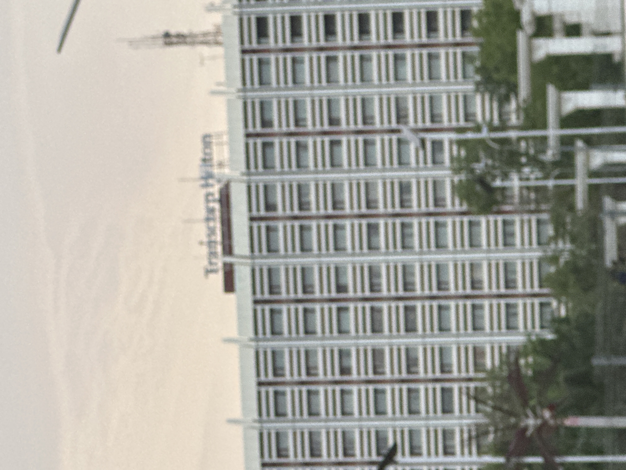 The image shows a tall building identified as the "Transcorp Hilton" in Abuja, Nigeria. The building is white with multiple rows of windows. Some trees are partially visible on the right side of the building. A crane can be seen in the distance at the top left of the image. The sky is overcast.