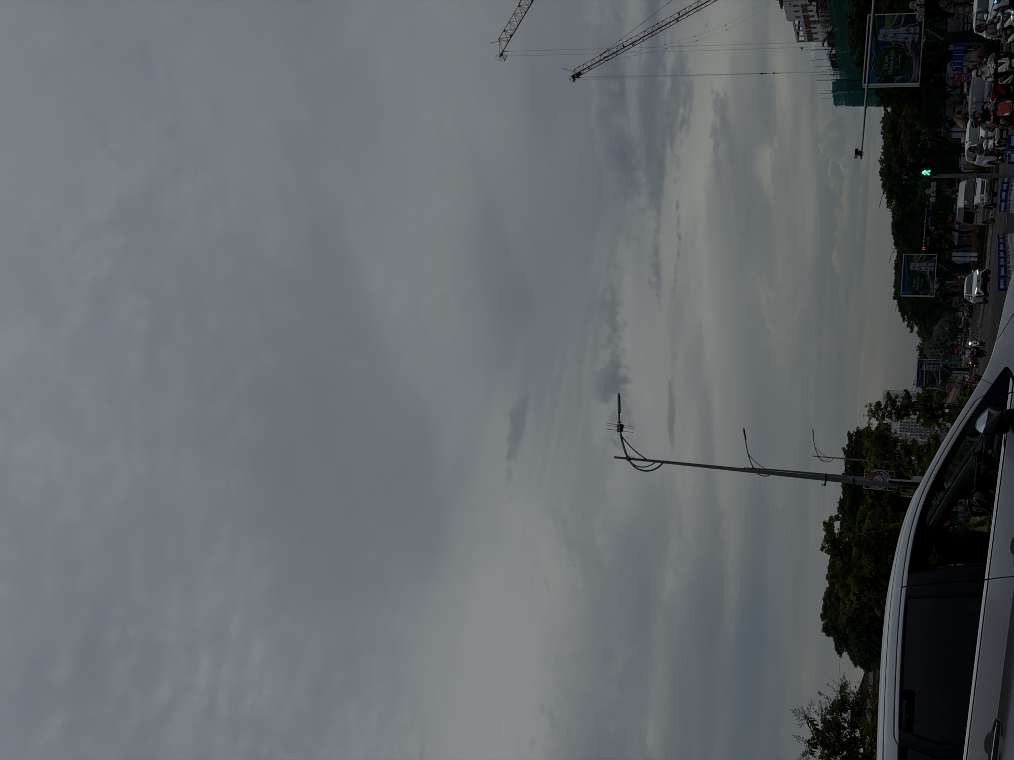 The image depicts a street scene in Đà Nẵng, Vietnam, under a cloudy sky. In the foreground, the left side of a silver vehicle is visible. The street is lined with trees, traffic lights, and several construction cranes in the background, indicating ongoing development. Various vehicles, including cars and motorcycles, are present on the road, with some people riding motorcycles. Billboards are also visible along the street. The overall atmosphere is overcast, suggesting possibly approaching rain or a generally gloomy day.