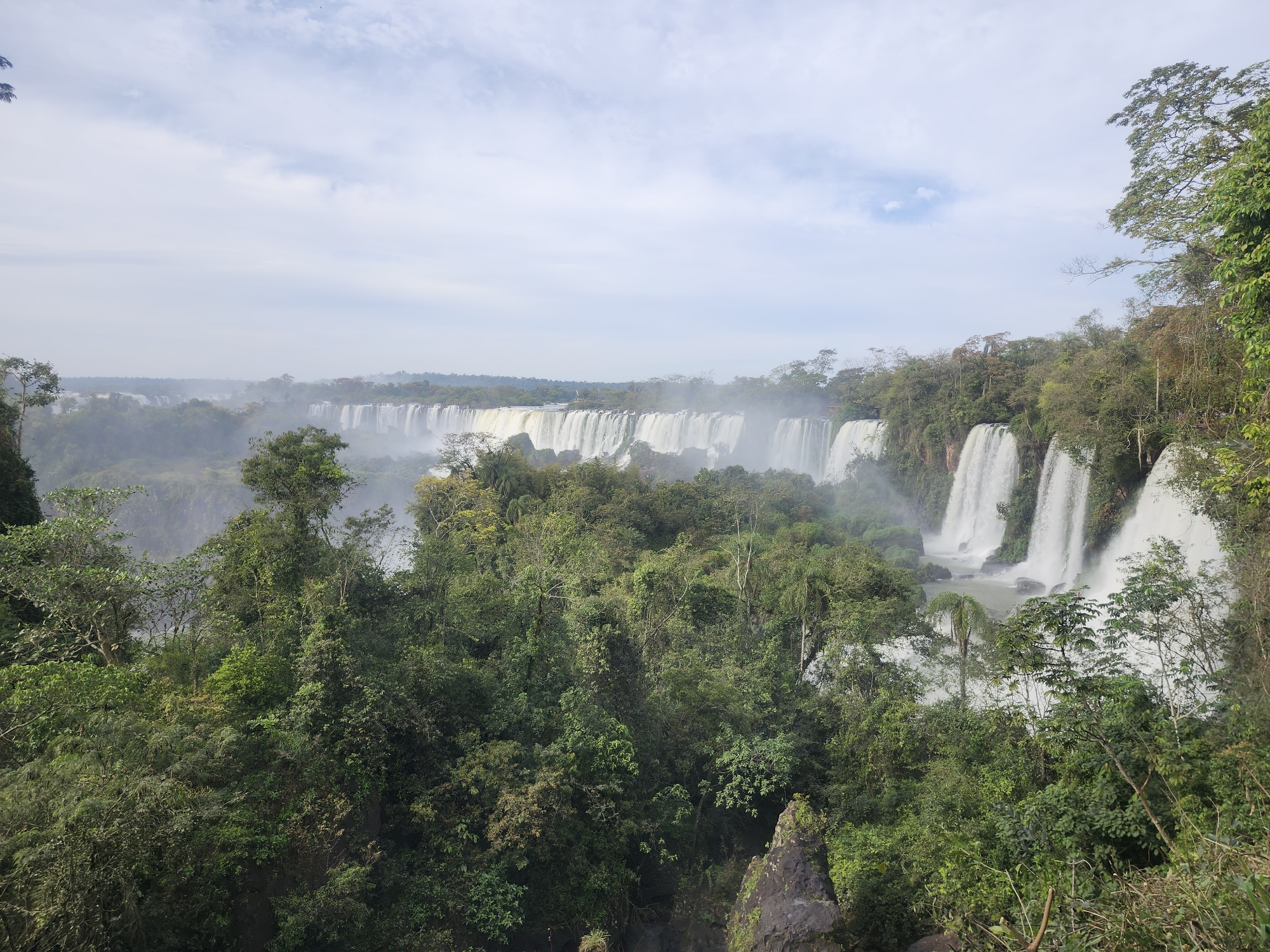 The image captures a panoramic view of Iguazu Falls, a natural wonder located on the border of Argentina and Brazil, near Puerto Iguazú, Argentina. The falls are a series of numerous individual waterfalls, with cascades of white water plunging into a misty abyss. Lush green rainforest foliage, dense with trees and vegetation, frames the foreground and extends towards the horizon. The sky above is partially cloudy with a pale blue hue, and a light mist hangs in the air, characteristic of the humid environment surrounding the falls. No people or man-made structures are immediately visible within the frame, emphasizing the raw, natural beauty of the landscape.