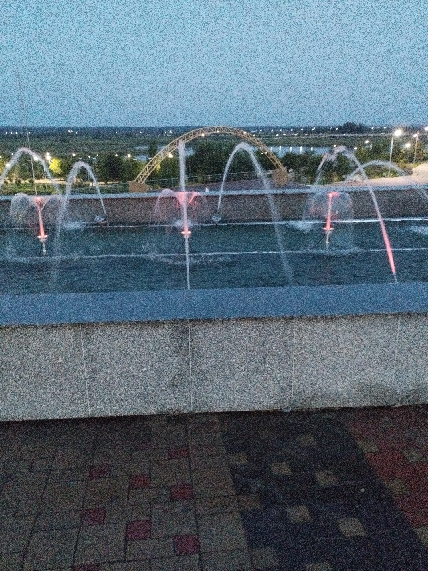 The image shows a fountain with several water jets, some of which are illuminated with pink light. The fountain is set against a backdrop of trees, a bridge, and a cityscape at dusk. The foreground consists of a patterned tiled walkway and a stone ledge. This fountain is located in Kostanay, Kazakhstan, and is part of the Fountain Complex on Dostyk Street. The image captures a serene evening ambiance with the water's movement and soft lighting.