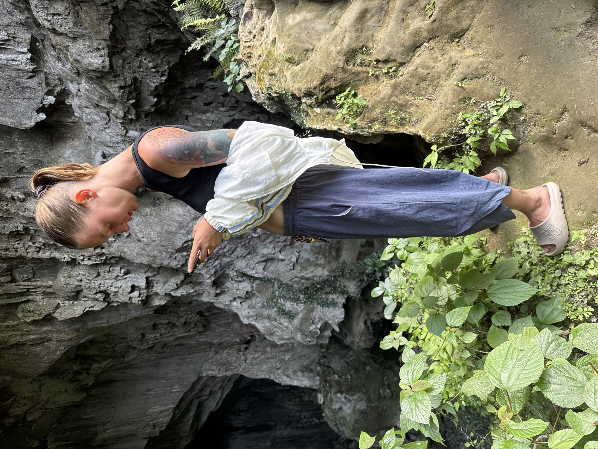 A woman with blonde hair tied in a ponytail is standing outdoors, looking at her phone. She is wearing a black tank top, a white jacket with blue stripes, and loose grey pants. She has tattoos on her right arm. The woman is standing near the entrance of a cave, with lush green foliage and rocky formations surrounding her. The scene appears to be in a natural environment, possibly Pù Luông Commune, Vietnam, known for its mountainous landscapes and caves.