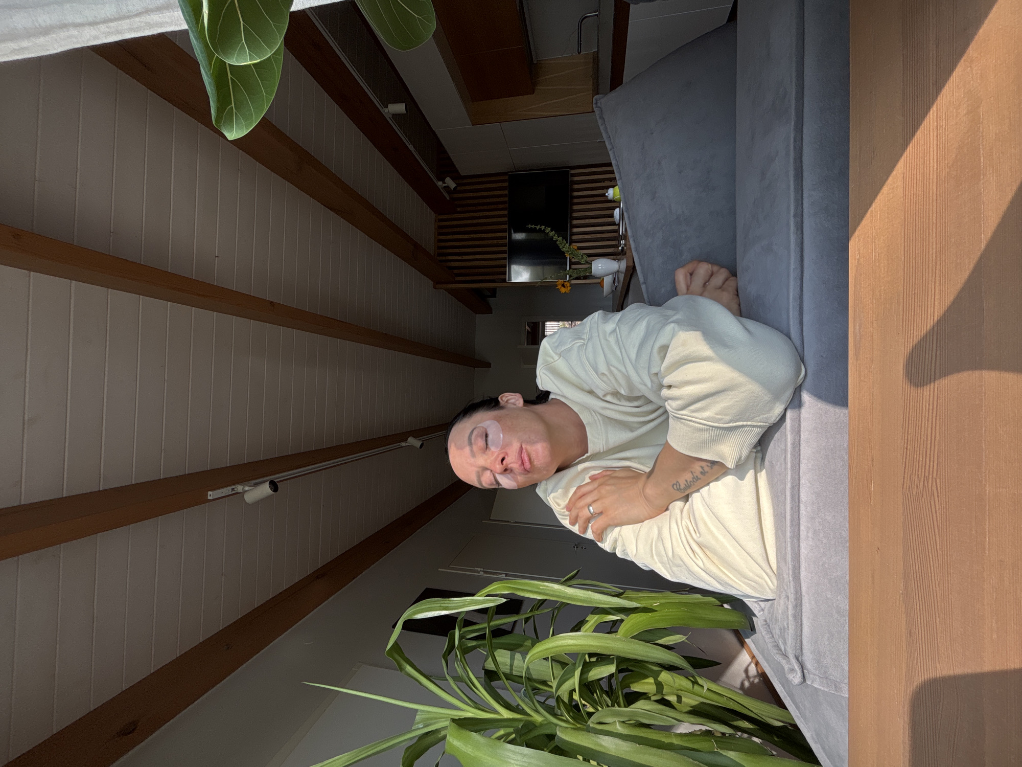 A person with eye masks lies on a gray sofa. The room has a sloped, beamed ceiling, a wall with horizontal wood slats, and plants. A television is mounted on the wall. The location and city are not determinable from the image.