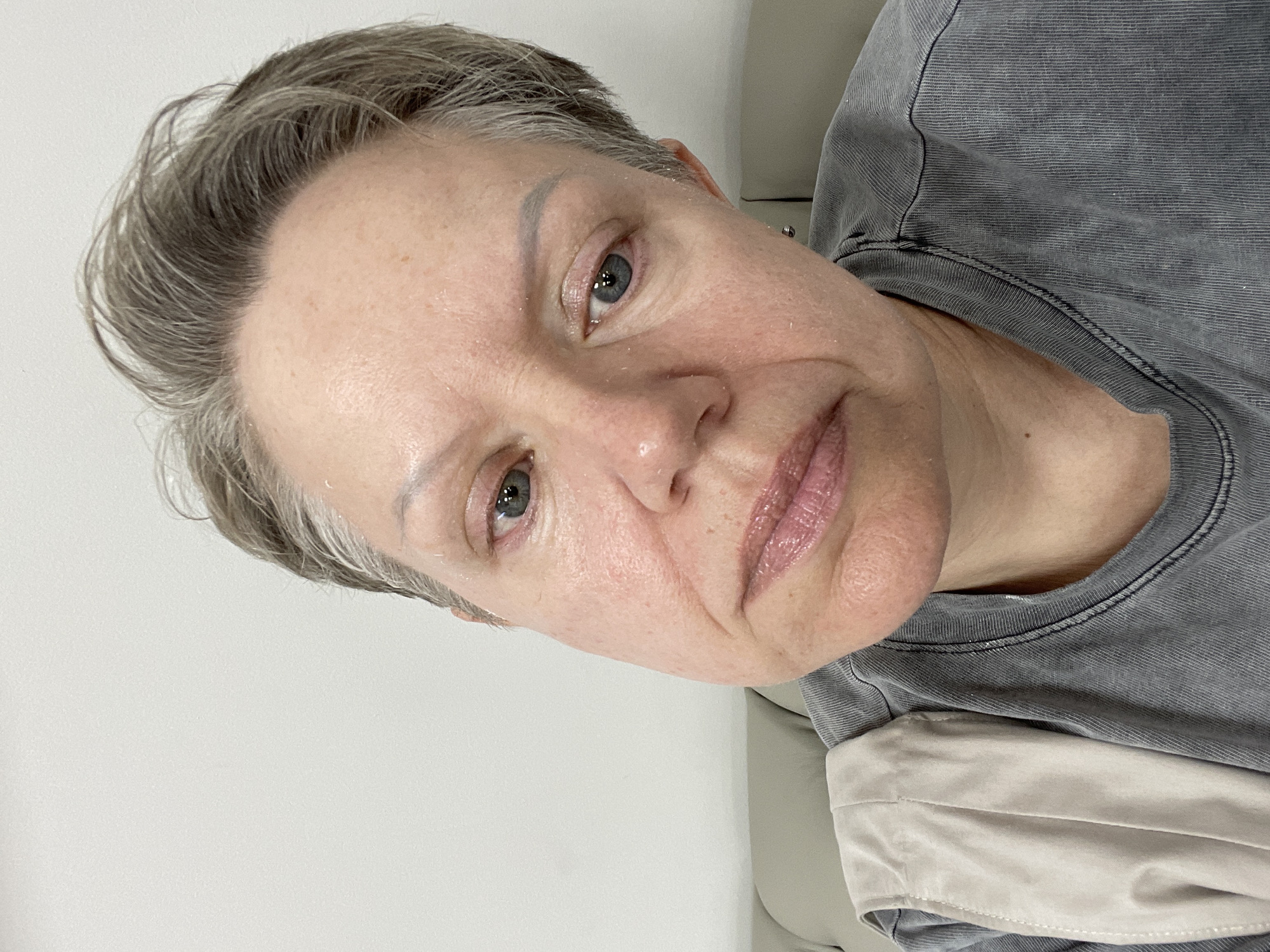 A close-up selfie shows a middle-aged woman with short, graying hair and blue eyes. She is wearing a gray t-shirt and a light-colored garment draped over her shoulder. Her face appears to have a light sheen, possibly from a skincare product. The background is a plain white wall, and a portion of a beige sofa is visible on the left side of the image. The lighting is even, suggesting an indoor setting. There is no discernible information in the image that places the subject in Almaty, Kazakhstan.