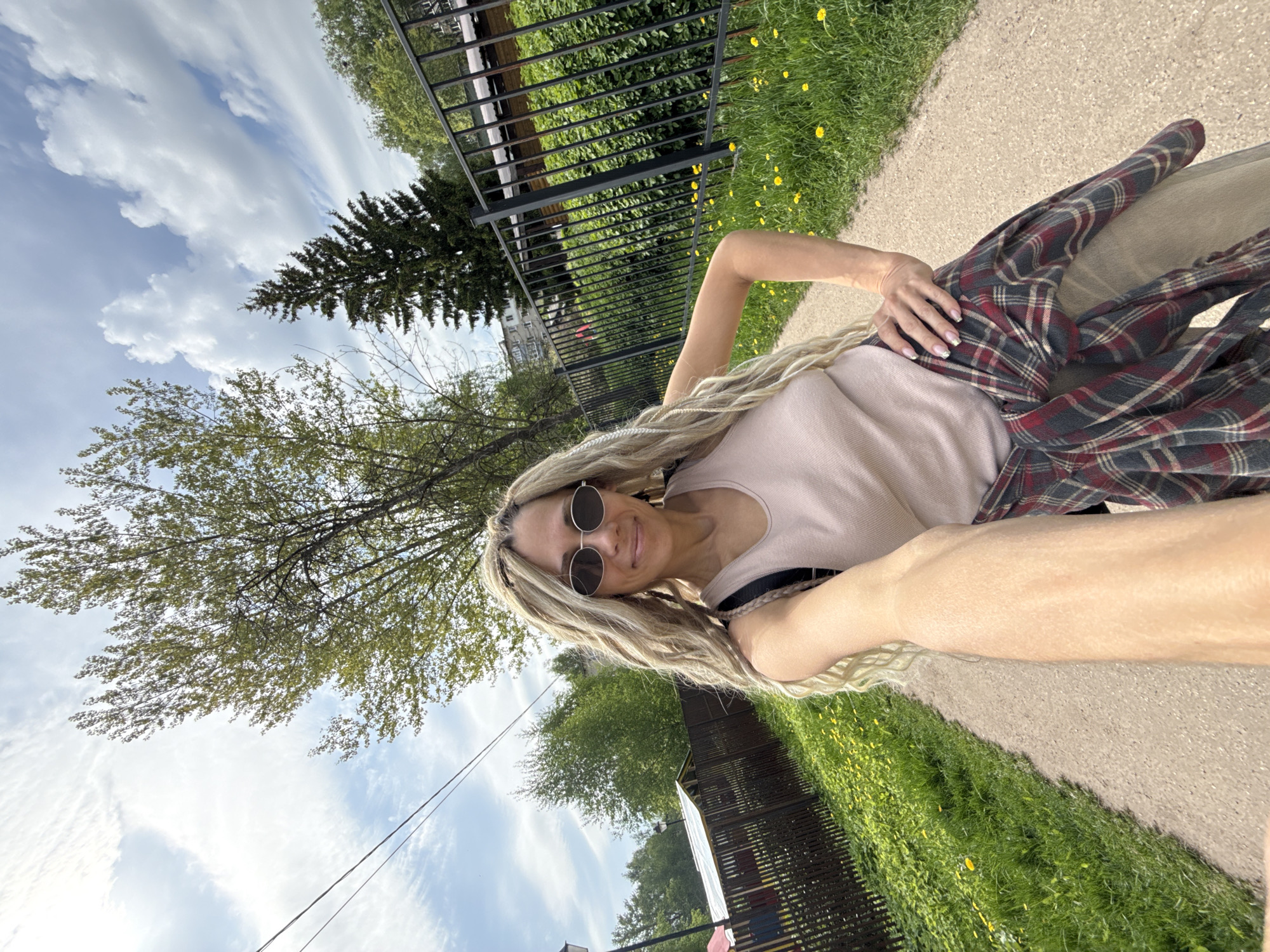 A woman with blond dreadlocks and round sunglasses takes a selfie on a paved path in Rusanovo, Russia, on a sunny day with scattered clouds. She wears a light pink top and a red and gray plaid shirt tied around her waist. A black metal fence borders the path on one side, with greenery visible behind it and yellow flowers in the grass. Trees are seen in the background against the cloudy sky. Overhead power lines are visible as well.