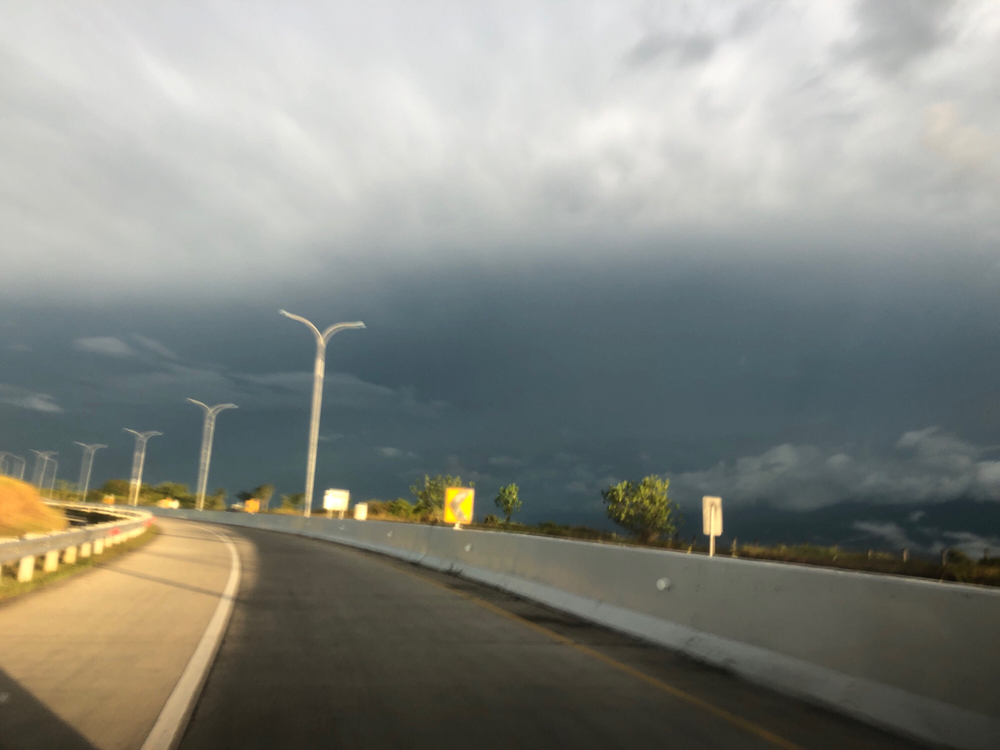 A motion-blurred, wide-angle shot captures a highway curving to the left under a dramatic, dark, and cloudy sky. The road is paved with asphalt, marked with a white line, and a lighter-colored lane to the left is also visible. A low concrete barrier separates the road from the shoulder and the grassy embankment. Multiple streetlights with a distinctive curved design are spaced along the highway. In the distance, a yellow traffic sign with a black arrow indicating a curve is visible, along with several trees and the suggestion of distant hills or mountains. The overall impression is one of an approaching storm. This scene is located in the city of Sihomlhok, Indonesia.