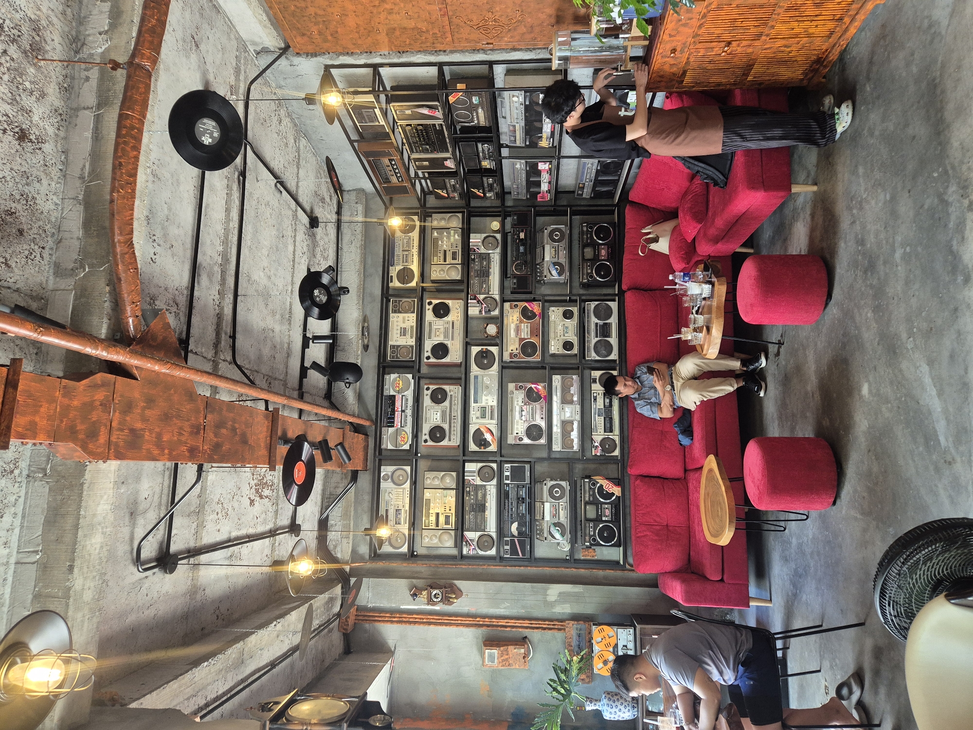 The image depicts the interior of a coffee shop in Da Nang, Vietnam, with a strong retro and industrial aesthetic. The walls are concrete with exposed pipes and orange-colored metal conduits. The ceiling also features exposed concrete and pipes. Decorative vinyl records are suspended from the ceiling.
The dominant feature of the cafe is a large wall unit filled with vintage boomboxes and other audio equipment. These are arranged on black metal shelves. In front of this wall is a bright red sectional sofa.
Two customers are seated on the sofa. One, a man wearing a grey t-shirt and dark shorts, is seated on the left side of the sofa and appears to be looking at something on the table in front of him. Another man, wearing a blue checkered shirt and light-colored pants, is seated on the right side of the sofa, looking at his phone.
A small, organically shaped wooden coffee table sits in front of the sofa, with a red ottoman beside it. Various glasses and drinks are on the table.
On the right side of the image, a barista, wearing a black t-shirt, brown apron, and striped pants, is seen preparing a drink behind a counter. The counter is made of distressed orange wood with a textured finish. A glass water dispenser and some plants are on the counter.
The floor is a polished concrete surface. A black electric fan is visible in the foreground, partially obscuring a light-colored chair. Other decorative elements in the cafe include a vintage clock on the wall and a vase with greenery. The overall atmosphere suggests a relaxed and trendy coffee shop environment.