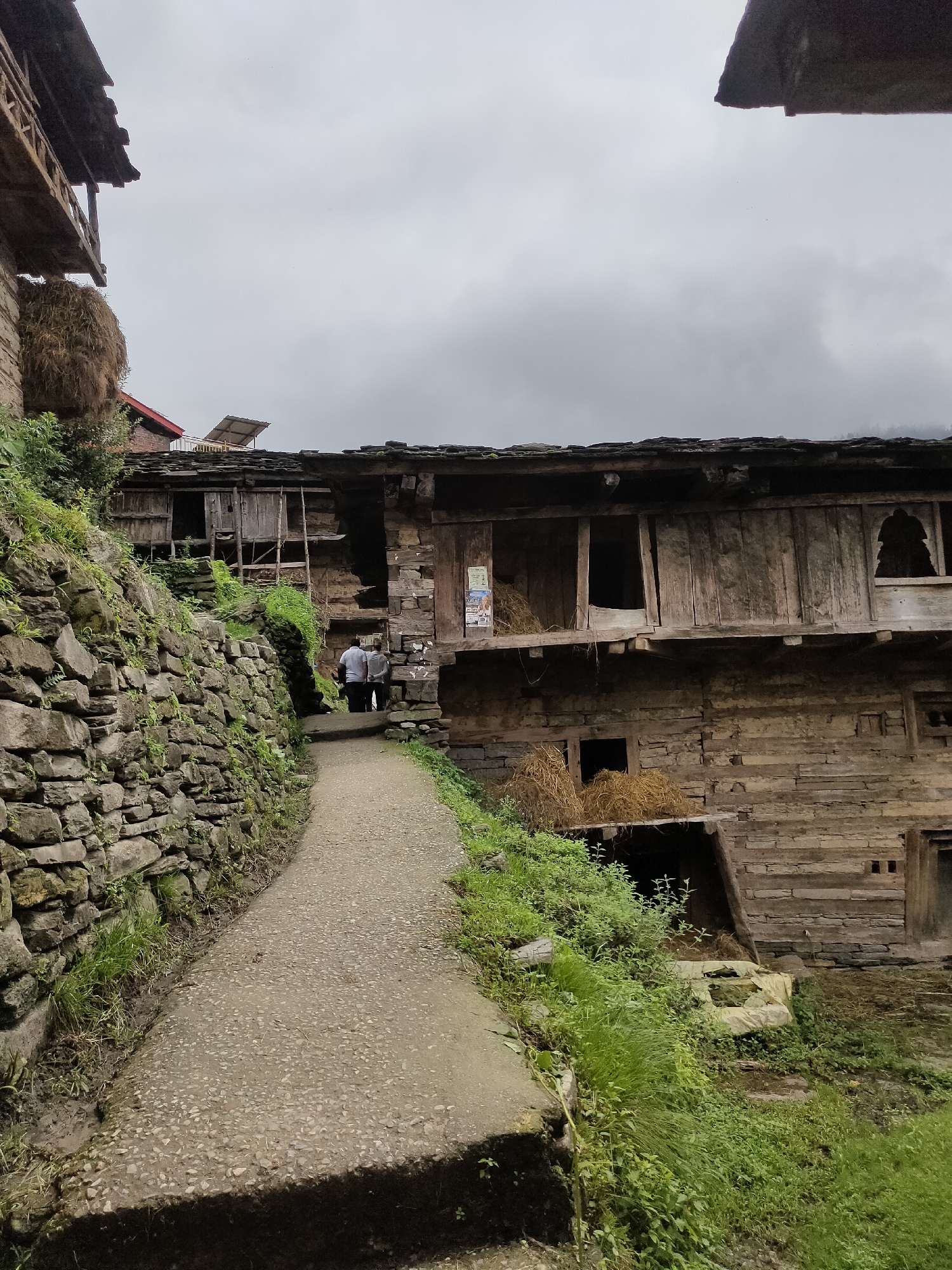 The image depicts a narrow, paved pathway curving uphill alongside a dry stone wall on the left. Overgrown green vegetation lines the path and the wall. On the right, a multi-story wooden structure with a dark, weathered facade is visible. The building appears to be constructed of timber planks and stone. Several openings, some with wooden shutters and others appearing as doorways or large windows, punctuate the structure. Bales of hay are stacked outside some of these openings. Two individuals wearing light-colored shirts are walking up the path towards the wooden building. The sky above is overcast with gray clouds. The scene is likely a rural setting in Damadi, India, given the architectural style and natural elements.