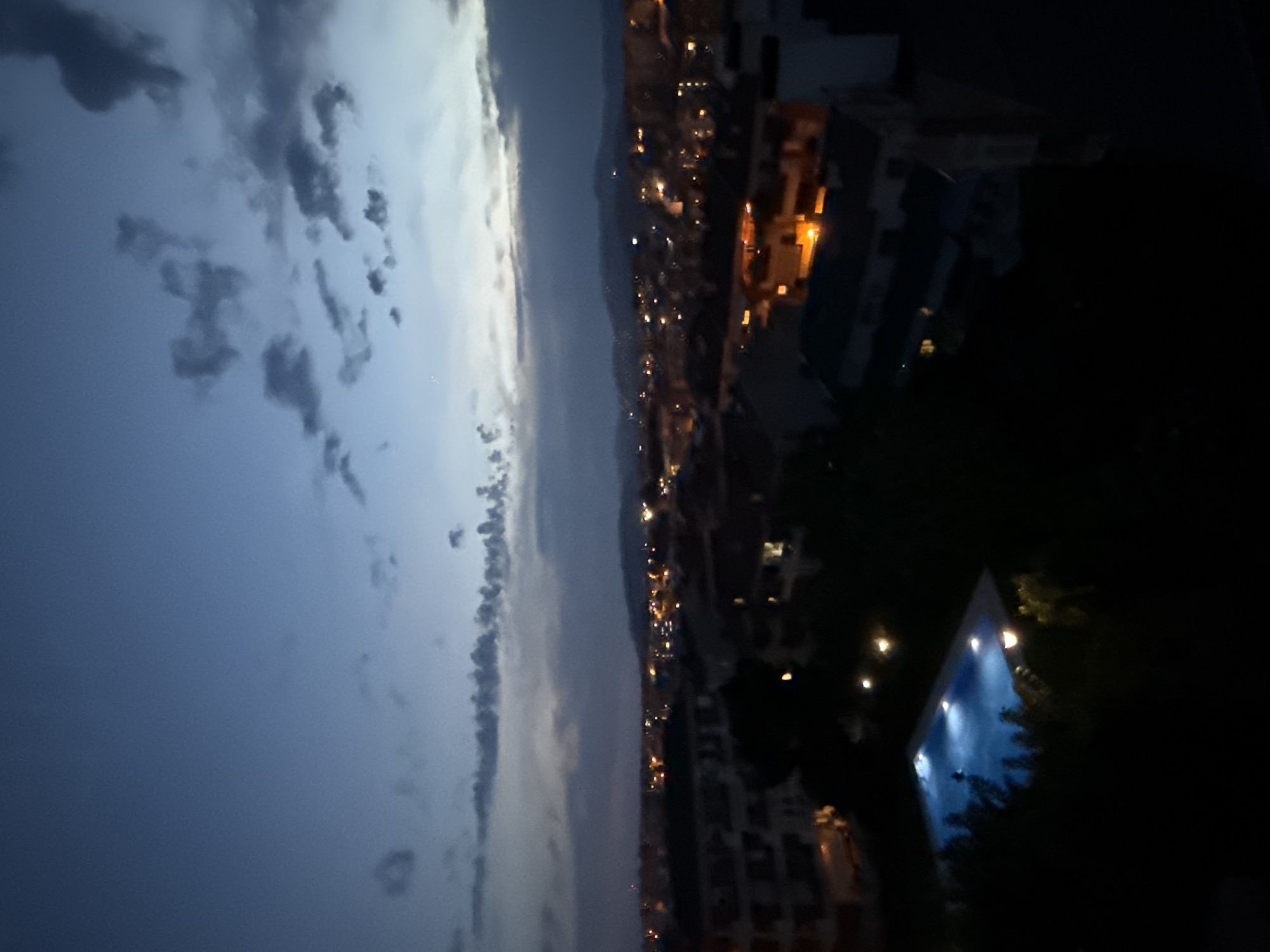 The image is a dusk or nighttime aerial view of a coastal city in Blanes, Spain. In the foreground, a rectangular swimming pool is illuminated with blue light, and a solitary figure can be seen swimming in it. Surrounding the pool are dark trees. Beyond the pool, the scene transitions to a view of a cityscape with numerous buildings and lights. The city extends towards a range of hills in the background. The sky above is a deep blue, transitioning to lighter hues near the horizon, with scattered clouds visible. The lights from the city indicate it is evening or night.