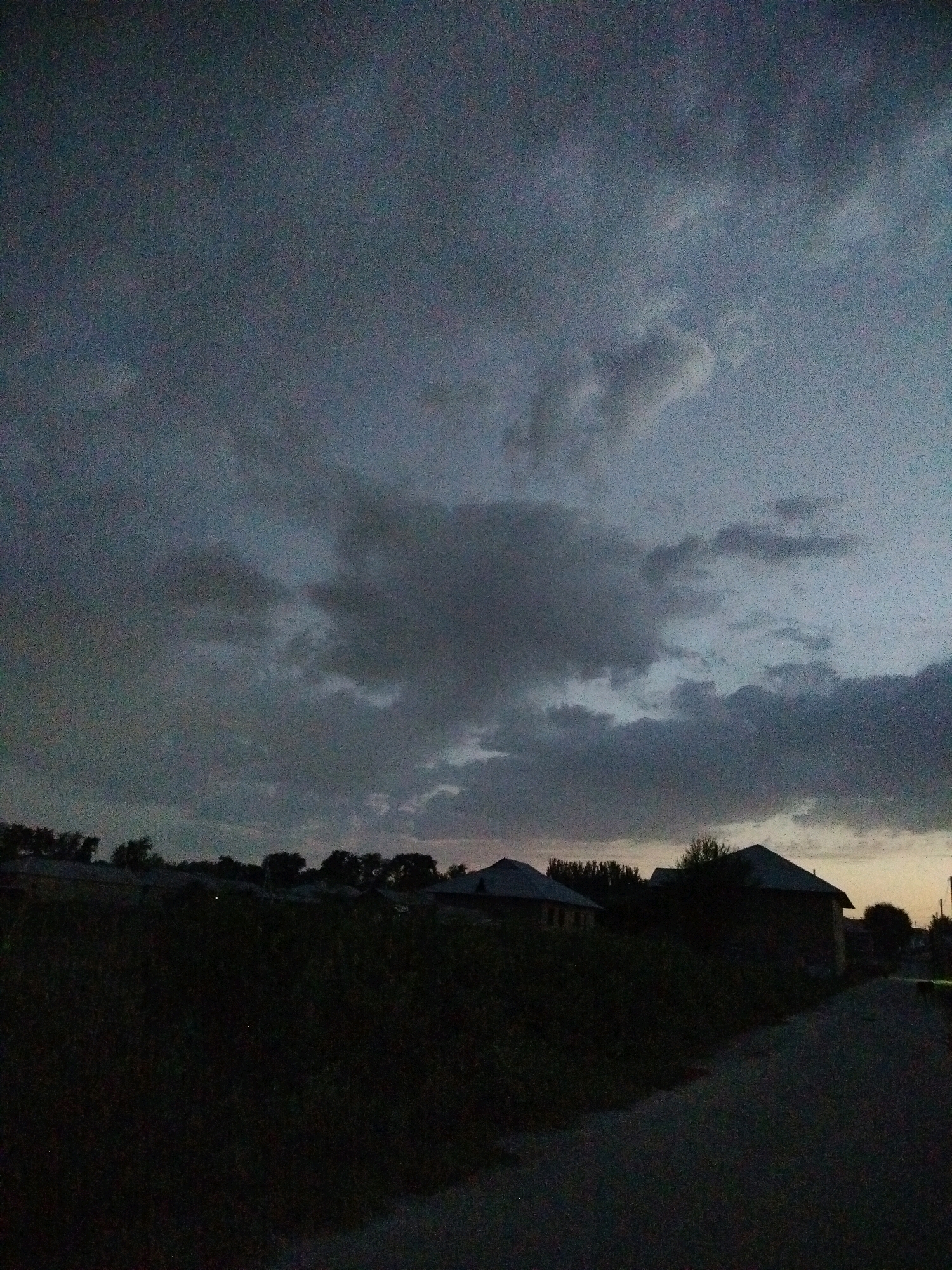 The image captures a dark, cloudy sky over a rural area at dusk, possibly in the city of Kölkent, Kazakhstan. The foreground is dominated by a dark, unlit road or path. To the left, a dense, dark patch of vegetation is visible, obscuring details of any structures beneath it. In the midground, silhouetted against the dim sky, are several houses with dark roofs. The sky itself is filled with heavy, dark clouds, with hints of lighter sky peeking through in the distance. There are no people or animals visible in the scene. The overall atmosphere is gloomy and still, characteristic of the period just before nightfall on a cloudy day.
