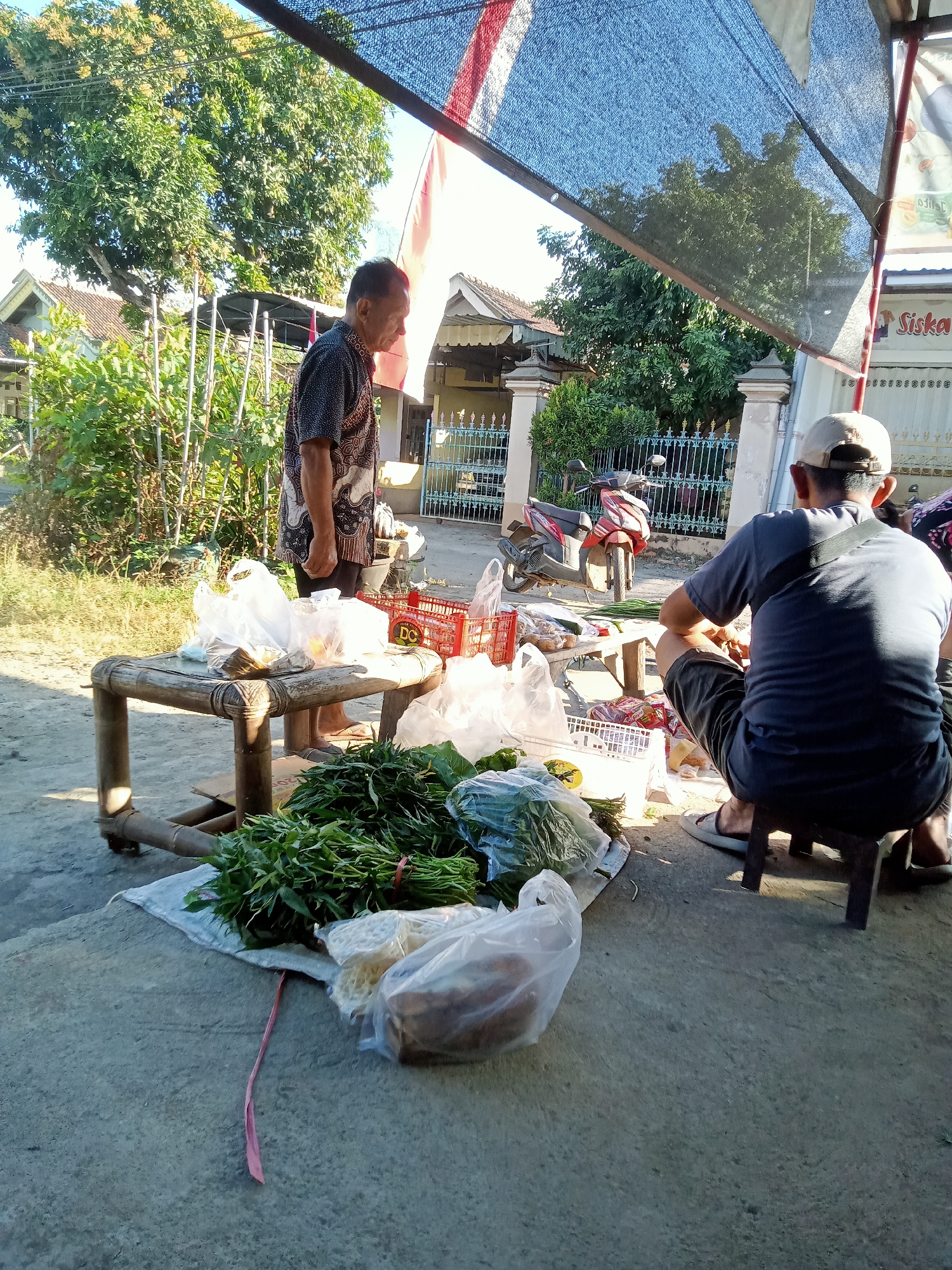 The image depicts an outdoor market scene in Simo, Indonesia. Two men are present. The first man, wearing a patterned short-sleeved shirt and dark pants, stands near a low bamboo table laden with plastic bags containing goods. He is facing away from the camera, looking towards the ground. The second man, wearing a grey t-shirt, dark shorts, and a baseball cap, is crouched on a stool, facing away from the camera and appearing to be engaged in an activity with his hands, possibly sorting or preparing items.
In the foreground, a variety of fresh produce, including leafy green vegetables bundled with red ties, are spread out on a white cloth. Several plastic bags containing other items are also visible, along with a red plastic crate. Behind the bamboo table, a motorcycle is parked. The background shows a residential area with houses, a fence, and lush green trees under a bright sky. A dark mesh canopy, possibly for shade, is stretched over part of the scene. The ground is a mix of concrete and dirt.