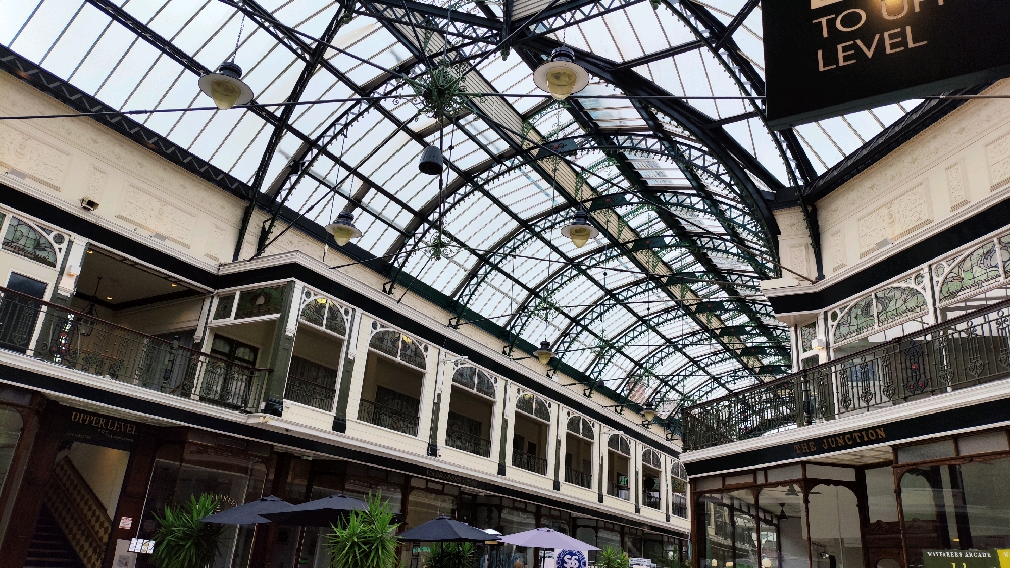 The image shows the interior of the Wayfarers Arcade in Southport, United Kingdom, a historic Victorian shopping arcade. The camera is positioned looking upwards, capturing the ornate glass and wrought-iron ceiling that arches over the walkway. Sunlight filters through the glass roof, illuminating the intricate metalwork and the interior architecture.
On either side of the arcade, two-story buildings with ornate facades and balconies line the walkway. The upper levels feature arched windows with decorative stained glass. The ground level has shop fronts, some with signage visible, including "UPPER LEVEL FOR THE HAIR LOUNGE" and "THE JUNCTION." Several black and grey umbrellas are set up on the ground, suggesting outdoor seating for a cafe or restaurant within the arcade. Lush green plants are also visible, adding to the atmosphere. Hanging lights are suspended from the ceiling structure. A black sign with white lettering reading "TO UP LEVEL" is visible hanging from the ceiling in the upper right portion of the image.
