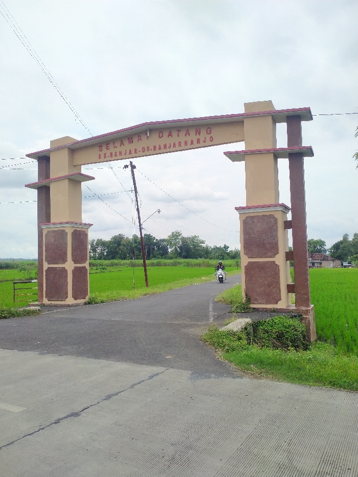The image captures a rural entrance marked by a decorative archway, likely in Kecamatan Kebakkramat, Indonesia. The archway spans a paved road, featuring beige pillars and a crossbeam adorned with reddish-brown accents. Prominently displayed on the archway is the text "SELAMAT DATANG" (Welcome) followed by "DK. BANJAR - DS. BANJARHARJO," identifying the hamlet (Dukuh) and village (Desa) being entered.
The scene is set under an overcast sky, suggesting a cloudy daytime. On both sides of the road, vibrant green rice fields stretch into the distance, indicating a fertile, agricultural landscape. A narrow asphalt road passes under the archway, leading towards a line of trees on the horizon. A single power pole stands near the left pillar of the arch, with power lines visible across the sky.
In the mid-ground, a person wearing a helmet is riding a white scooter or motorcycle away from the viewer, having just passed or in the process of passing under the archway. The ground in front of the archway is a lighter concrete slab, transitioning to the darker asphalt road. The overall impression is that of a quiet, welcoming entrance to a village surrounded by rice paddies.