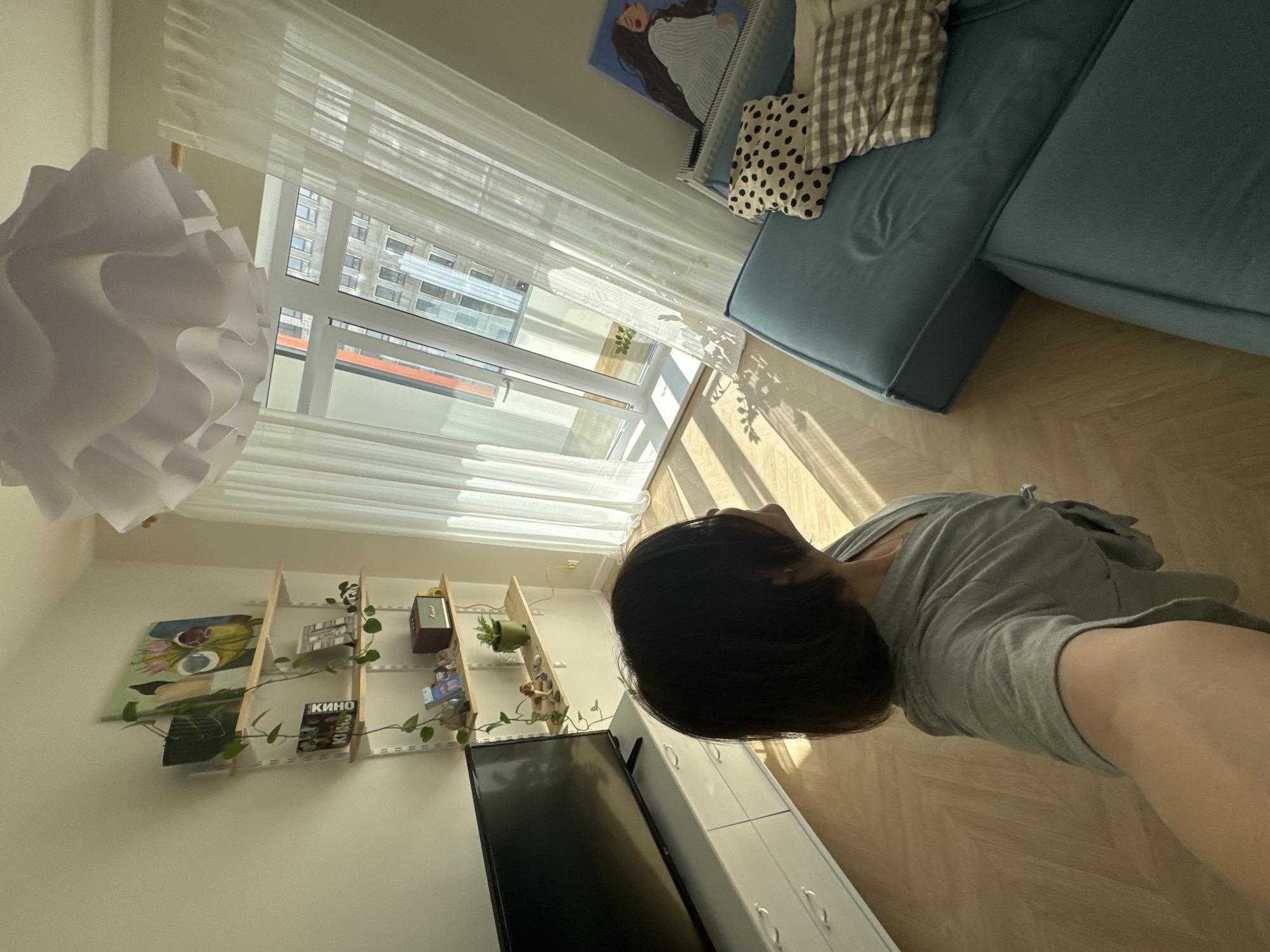 A person with short black hair is shown from the shoulders up, wearing a gray T-shirt. The scene includes a room with a blue sofa with patterned pillows, a large window with sheer curtains, a white pendant lamp, and a wooden herringbone pattern floor. Against the wall are shelves with various items including plants, paintings, and a black television on top of white drawers. The view from the window suggests an urban setting with buildings, possibly located in Izhevsk, Russia, though this is not definitively provable within the image.