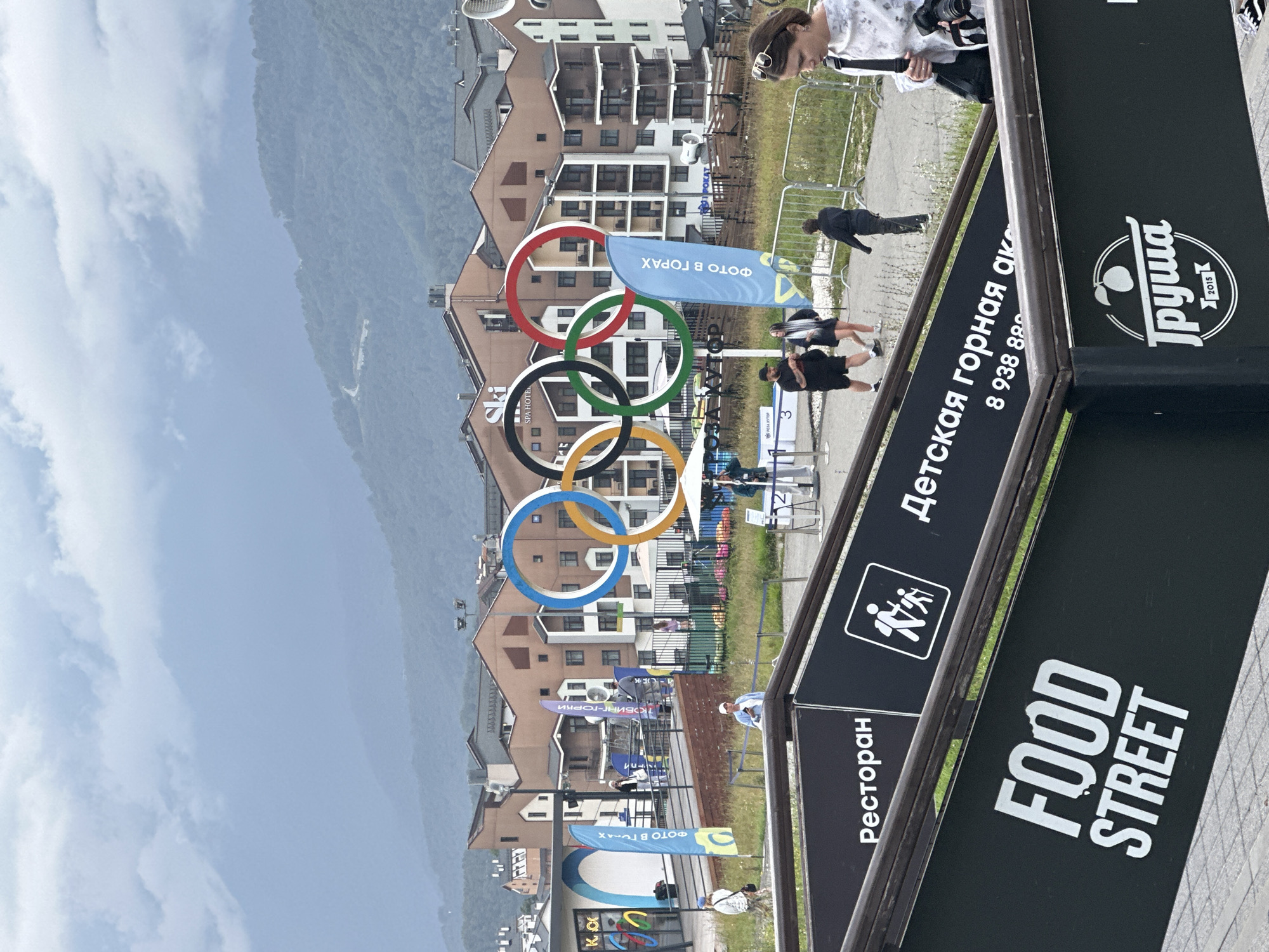 In Estosadok, Russia, a scene unfolds with the iconic Olympic rings prominently displayed in the background, set against a backdrop of multi-story buildings and misty mountains. The Olympic rings, a symbol of sports and international competition, stand tall, drawing attention to the resort town. In the foreground, a woman with sunglasses perched on her head walks by, carrying a camera, hinting at a tourist or photographer capturing the sights. Another woman, holding a black bag and a camera, is also visible, suggesting they are both part of a group enjoying their time in the picturesque location. Signs in the foreground provide further information about the offerings, with "FOOD STREET" and "Ресторан" (Restaurant) indicating dining options. A phone number, "8 938 88," is also visible, likely for local services. The environment appears to be a public space, possibly a promenade or entrance to a recreational area, with pathways and railings guiding visitors. The presence of these elements creates a dynamic scene, capturing the essence of a popular tourist destination.