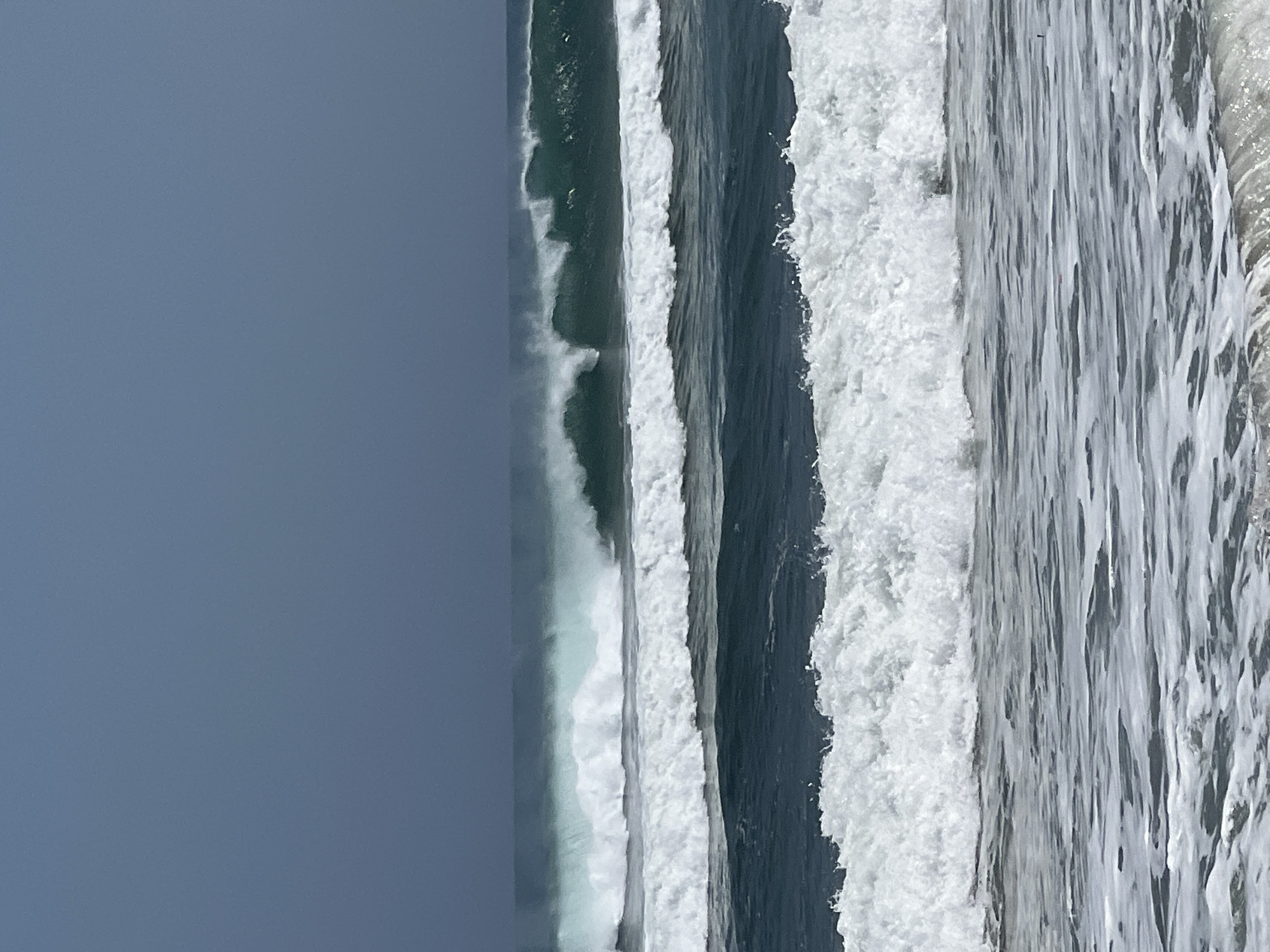 The image depicts a seascape with multiple waves breaking in the ocean. The sky is a deep, overcast blue. In the foreground, the crest of a wave is breaking with white foam. Behind this, a larger wave with a more defined peak is rolling in. Further out, another series of waves are breaking, with the largest wave showing a hollow tube formation, indicating a powerful swell. The water color transitions from a dark blue in the mid-ground to a lighter, foamy white in the foreground. The scene is characteristic of the popular surf break at Kuta Beach, Bali, Indonesia, known for its consistent waves. No people or man-made structures are visible in this frame.