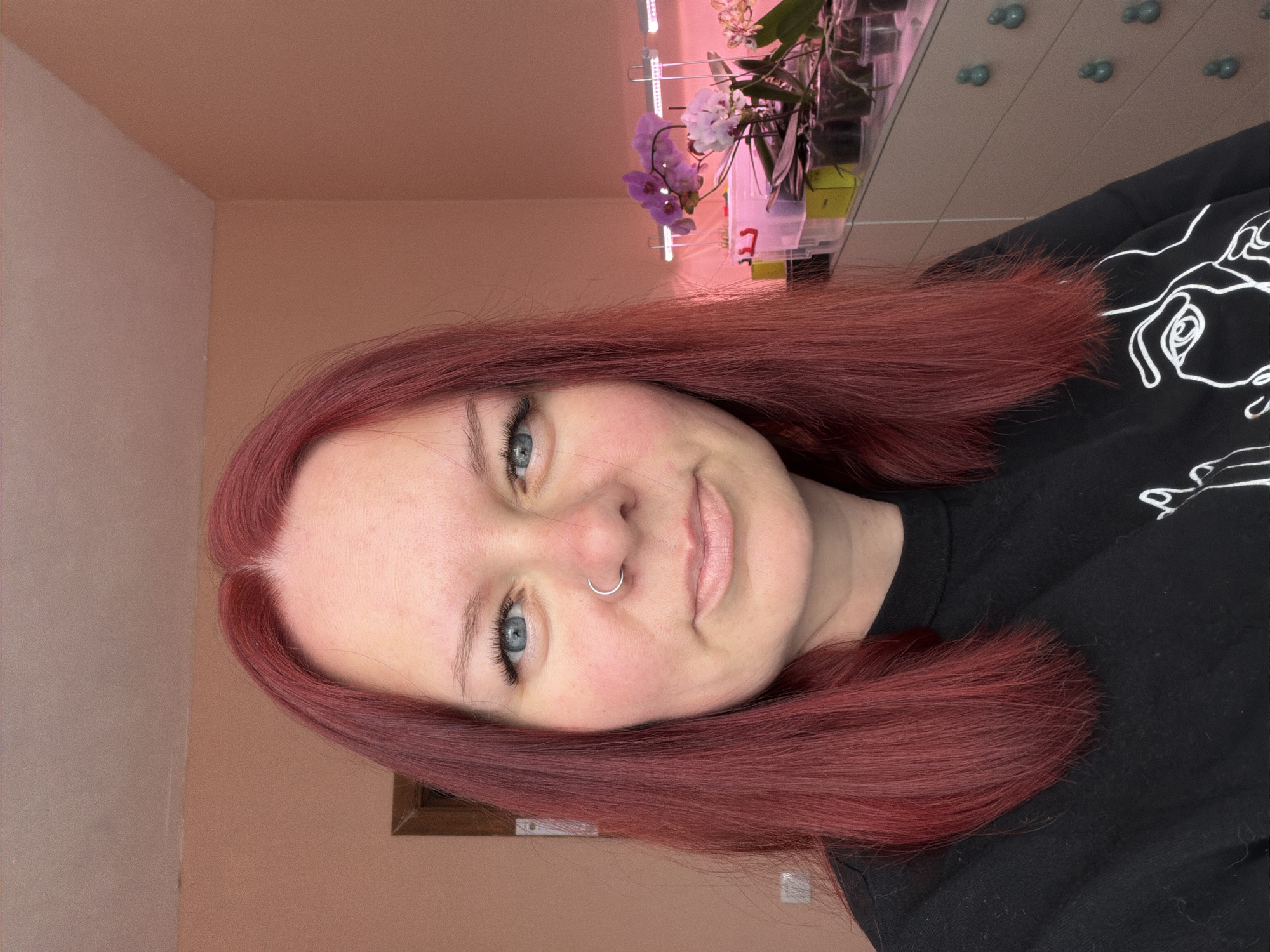 An adult female is depicted in a close-up indoor portrait, presumed to be in the city of Chaikovsky, Russia. She has medium-length, straight, red hair parted slightly off-center. Her light blue eyes are open, and she wears a silver hoop nose ring in her left nostril. She exhibits a neutral expression with a slight smile, looking directly at the viewer. She is wearing a black long-sleeved shirt or sweatshirt with a white, abstract line-art design partially visible on the lower right. The background features light peach-colored walls. To the left, a portion of a wooden door frame is visible. To the right, a multi-drawer dresser or cabinet with distinct light blue spherical knobs holds several potted plants, predominantly orchids with purple and white blooms. Above the plants, two horizontal LED grow lights emit a pinkish-purple glow. Other containers, including a white bin with a red character, are also present on the surface.