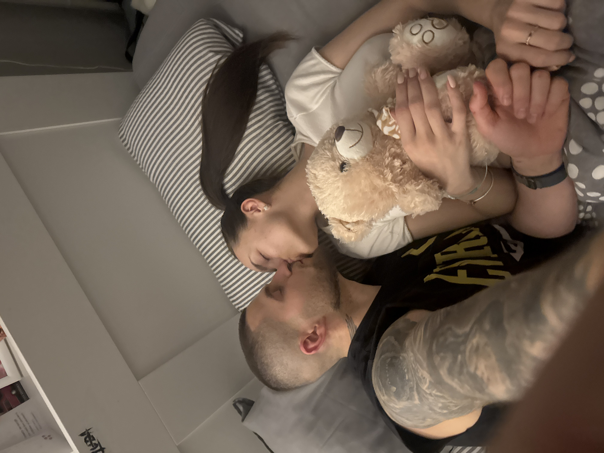 A couple is kissing on a bed. The woman is lying on a striped pillow, holding a teddy bear, and wearing a white shirt. The man is wearing a black shirt with yellow writing and has a large tattoo on his arm. The location is a room with a gray wall and a bookshelf in the bottom left corner. There is no indication of the location being in Sofia, Bulgaria.