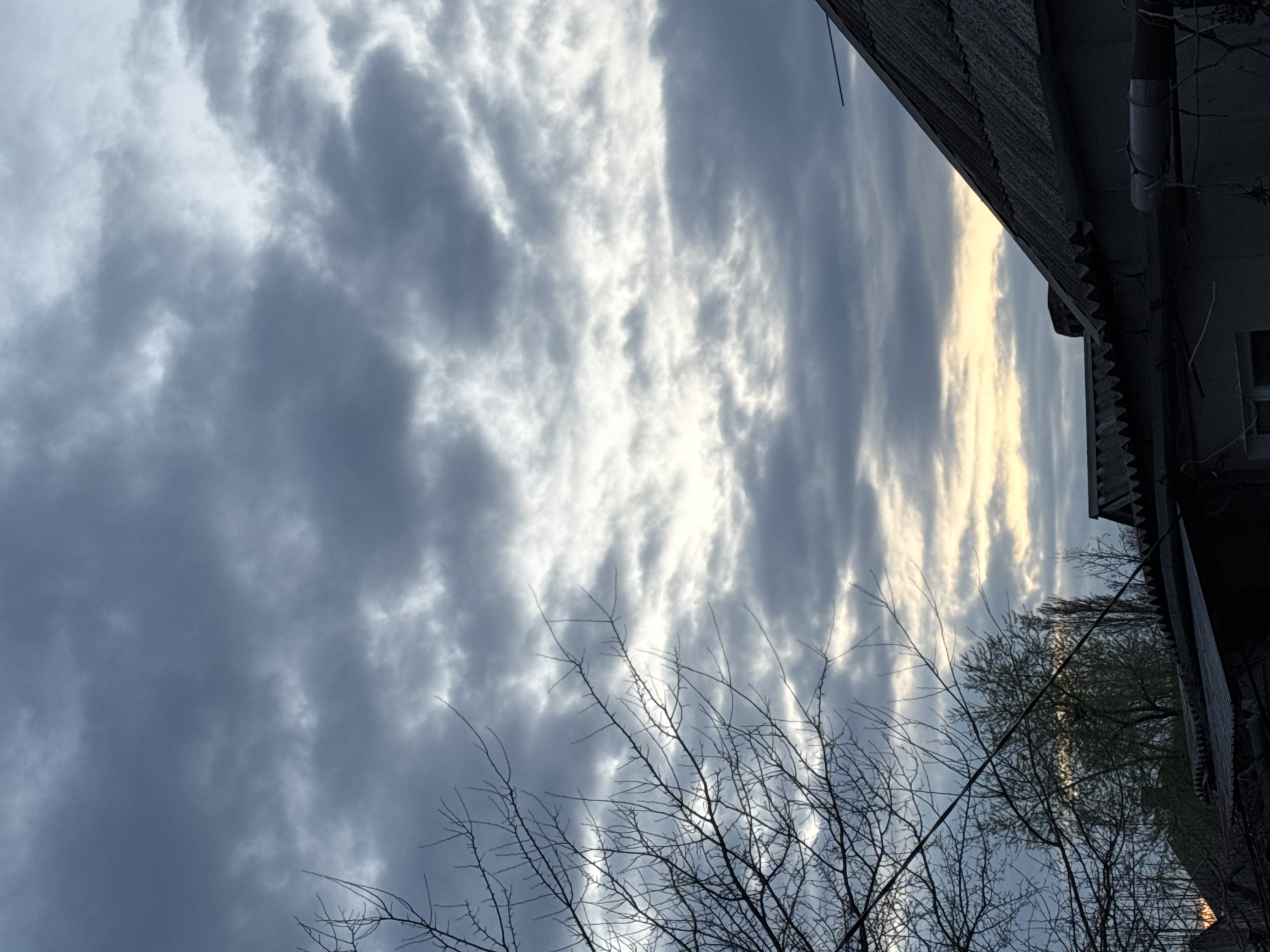 The image depicts a cloudy sky with bare branches and trees in the foreground. A building's edge is visible at the top right corner. The location is likely urban, suggested by the building, but the specific city is unknown. The clouds are a mix of dark grey and lighter, potentially sunlit patches. The overall environment suggests either late autumn or early spring due to the absence of leaves on the branches.