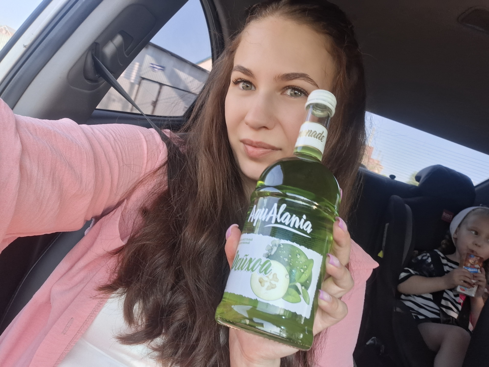 The image shows a young woman in a car, holding a green bottle of "Aqua Alania Feijoa" lemonade. She is wearing a pink shirt. A child is visible in a car seat behind her, drinking from a juice box. The background suggests an urban environment. The location is Chaikovsky, Russia.