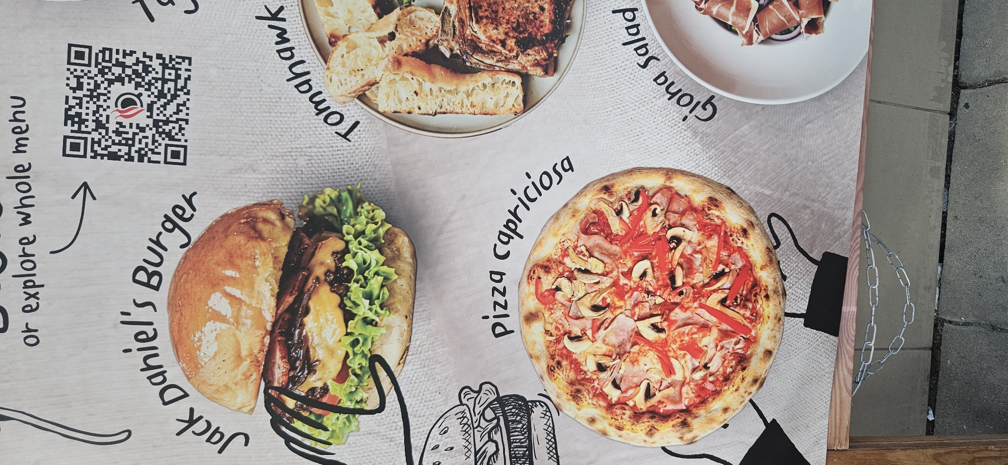 The image displays a menu board for a restaurant, featuring various food items. In the top left, a handwritten arrow points to a QR code, with text above reading "or explore whole menu". Below this, a large burger is pictured, labeled "Jack Daniel's Burger". To the right of the burger, a steak and bread are shown, labeled "Tomahawk". In the lower half of the image, a pizza is prominently displayed with the label "Pizza capricciosa". Further to the right, a salad is partially visible in a bowl, labeled "Giovanna Salad". The menu board is set against a textured background, possibly a wall or a canvas. In the foreground, part of a wooden structure and metal chains are visible, suggesting the menu board is part of an outdoor establishment or a temporary display. There are no people visible in the image. The text and images are clear and well-lit, indicating the photograph was taken in daylight. The specific location in Krakow, Poland, cannot be determined from the visual information provided in the image.