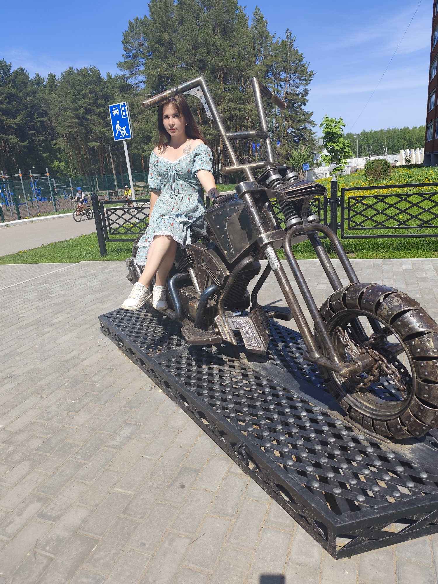 A young woman is sitting on a metallic motorcycle sculpture located on a paved area in Chaikovsky, Russia. She is wearing a short, light-blue dress with floral patterns and white sneakers. The motorcycle is intricately crafted from metal, featuring a large front tire and a prominent handlebar design. The motorcycle is positioned on a black, metal grate platform. Behind the woman and the motorcycle, there is a playground with a blue sign displaying pedestrian crossing with wheelchair. A fence separates the paved area from a grassy area with trees. Two children are riding bicycles in the background, and a multi-story building is partially visible to the right of the scene. The sky is clear and blue.