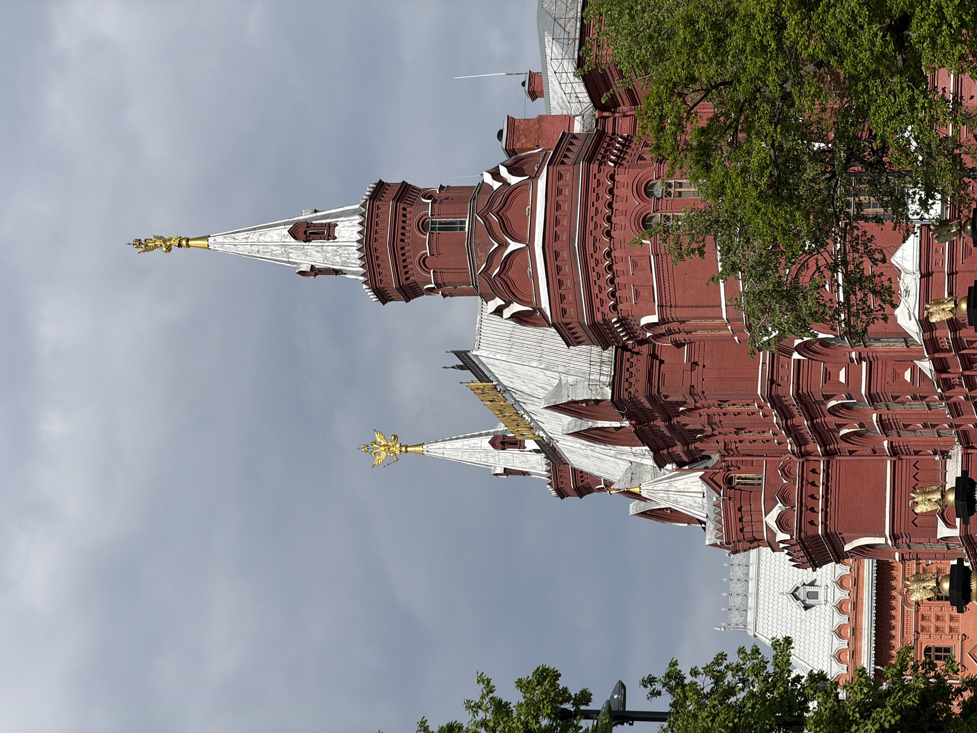 The image depicts the Moscow Kremlin in Russia, showcasing its distinctive red brick walls and towers. The architecture features a blend of medieval and Renaissance styles, characterized by pointed spires topped with golden eagles, ornate detailing, and white accents that contrast with the red brick. The sky is overcast, creating a soft, diffused light that highlights the textures of the building. Trees with green foliage are visible in the foreground and partially obscure some sections of the Kremlin, adding a natural element to the urban landscape.