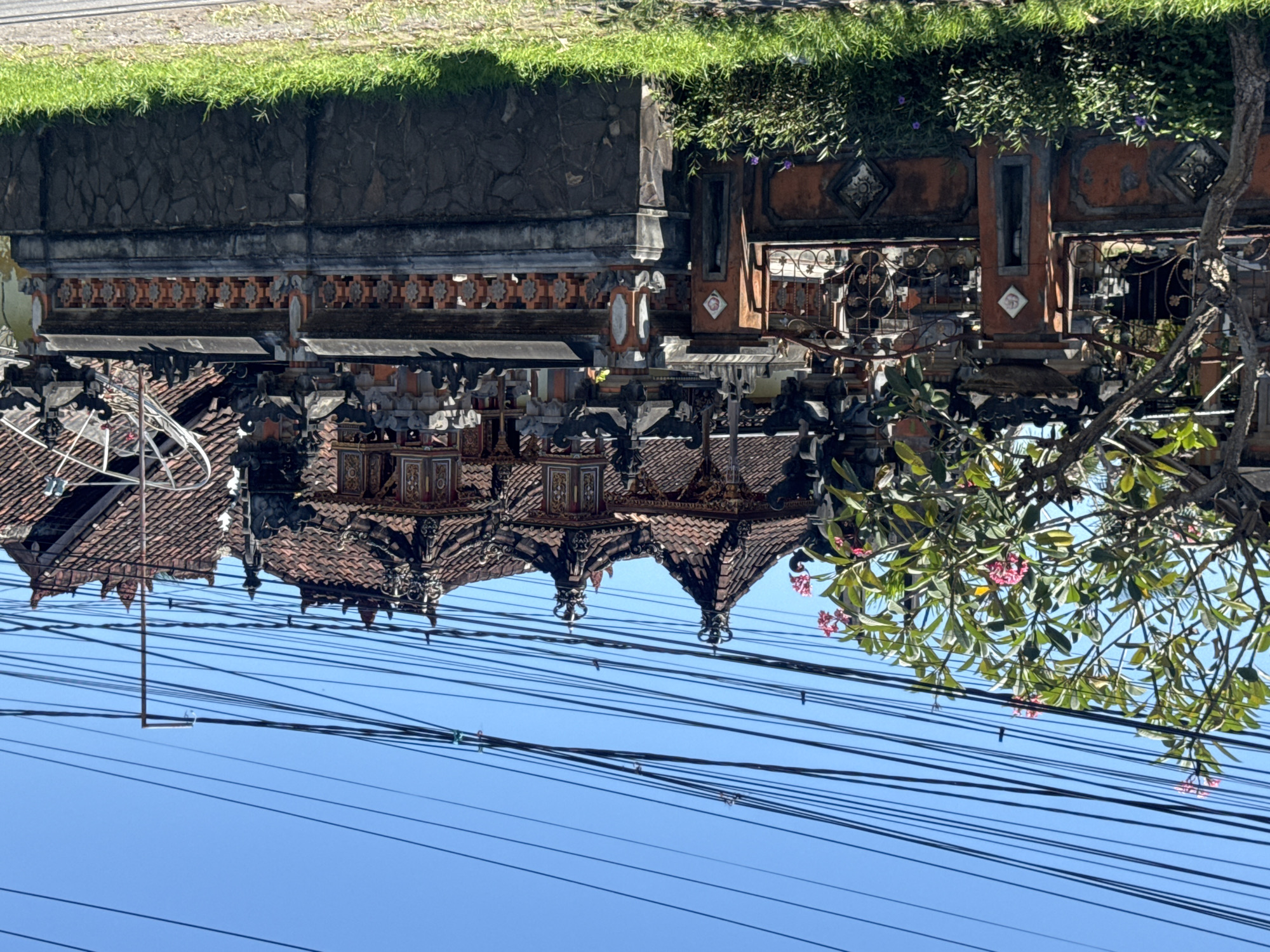 The image shows a low-angle view of Balinese architecture against a bright blue sky filled with utility wires. The architecture features tiered roofs with detailed decorations. A tree with pink flowers is visible on the right. Above the architecture is a wall topped with grass and plants. This image was taken in Kubu, Indonesia.