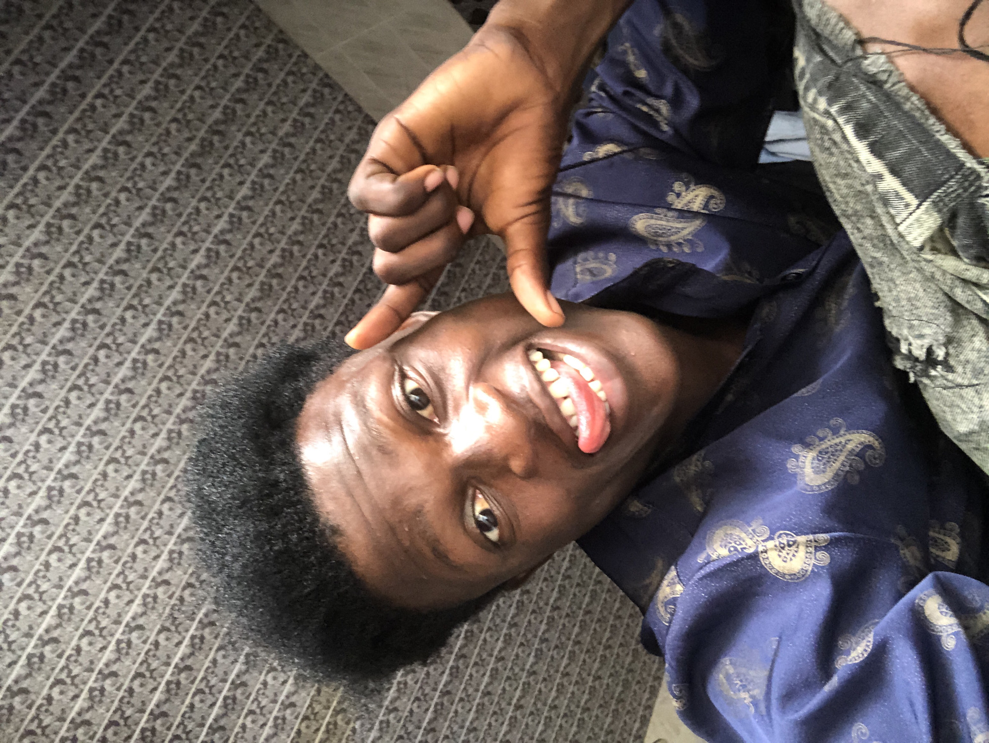 A young Black male, appearing to be in his late teens or early twenties, is captured in a close-up selfie taken indoors. He has dark, curly hair and is wearing a dark blue shirt with a gold paisley pattern. His head is tilted to the side, and he is sticking his tongue out with his mouth open in a wide smile, revealing his teeth. His right hand is raised, with his index finger and thumb forming a "L" shape near his cheek, while his other fingers are curled inwards. The background appears to be a patterned ceiling with diagonal lines and a textured gray surface. The lighting suggests an indoor setting with a light source from above and slightly to the left, creating highlights on his face and shoulders. The overall impression is one of playfulness and a casual demeanor. The location is identified as Ajah, Nigeria, implying this is a personal photograph taken in a residential or public space within that area.
