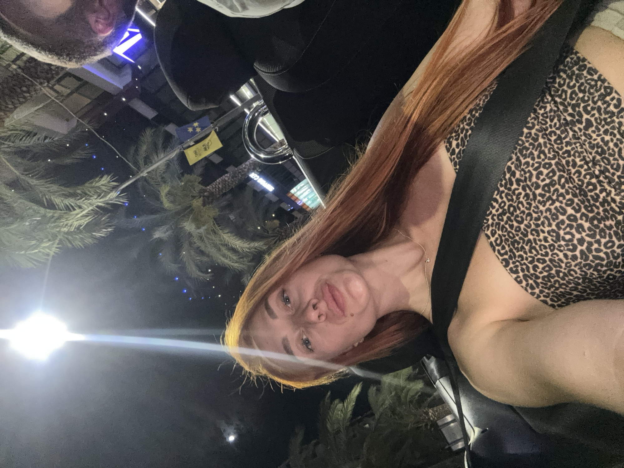 An outdoor night-time selfie, taken from the perspective of the driver's seat of a convertible, shows a young woman with long, reddish-blonde hair. She is wearing a leopard-print bandeau top and a silver necklace. A black seatbelt is diagonally across her chest. Her head is slightly tilted, and her lips are pursed. In the background, palm trees and illuminated buildings are visible, suggesting a location in Budva, Montenegro. A bright overhead light casts a glare on the scene, and part of another person's head is visible in the upper right corner of the frame.
