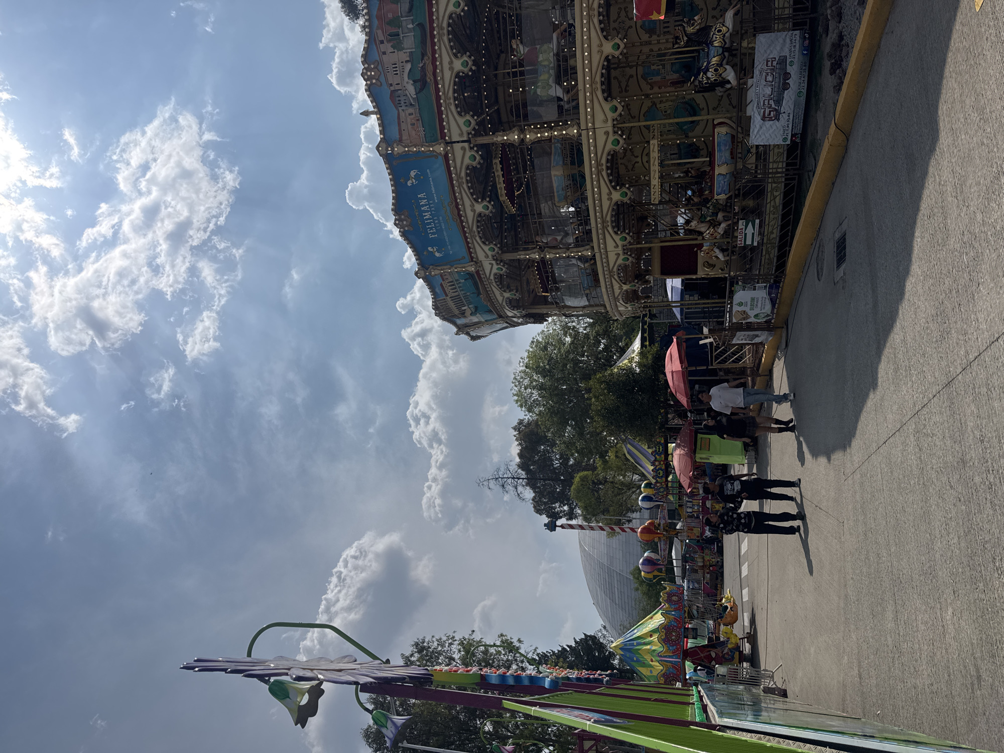 The image shows an outdoor scene in the city of Puebla, Mexico, featuring an amusement park setting. In the background, a multi-tiered carousel with decorative painted panels and ornate detailing is visible. Several individuals are walking along a paved path adjacent to the carousel. One person holds a red umbrella. A large dome-shaped structure can be seen behind trees in the distance. An amusement park ride with colorful decorations is in the foreground. The sky is partially cloudy.