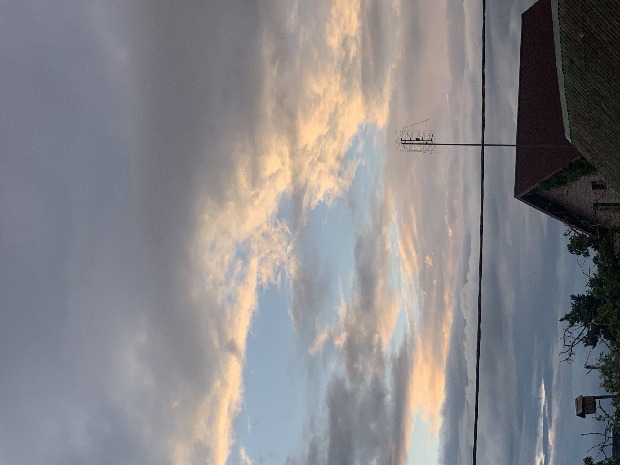 The image shows a view of the sky above Kapyĺ, Belarus, during what appears to be either sunrise or sunset. The sky is filled with a mix of dark, grey clouds and lighter clouds illuminated with warm, peachy tones. A clear patch of pale blue sky is visible in the upper center. In the foreground, a black wire stretches horizontally across the frame. Below the wire, the rooftops of several buildings are visible, including a red-tiled roof with ivy growing on a brick wall beneath it, and another corrugated roof. A tall antenna stands on a pole above the red-tiled roof, and a small birdhouse is perched on a pole to the left of the frame. Trees and foliage are present at the bottom of the image.