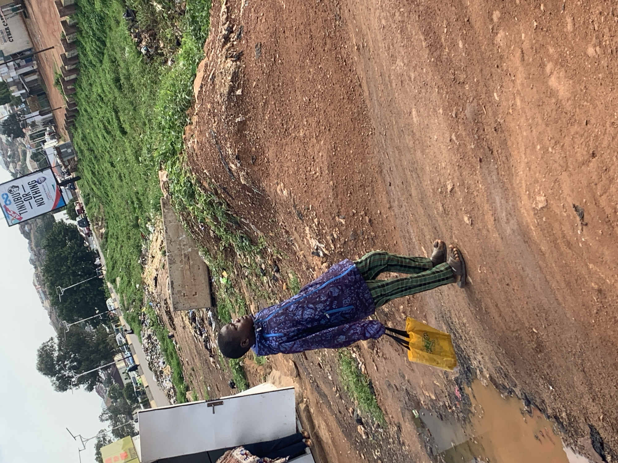 In Gura Top, Nigeria, a young boy walks along a dirt path next to a pile of garbage. He is wearing a blue patterned jacket and green checkered pants, and he carries a yellow bag in his right hand. In the background, a white door is visible, along with other structures and a grassy area dotted with trees. A billboard advertising "Tinubu - Or Nothing" stands near the road, which is lined with vehicles. The overall scene depicts a somewhat unkempt urban environment.