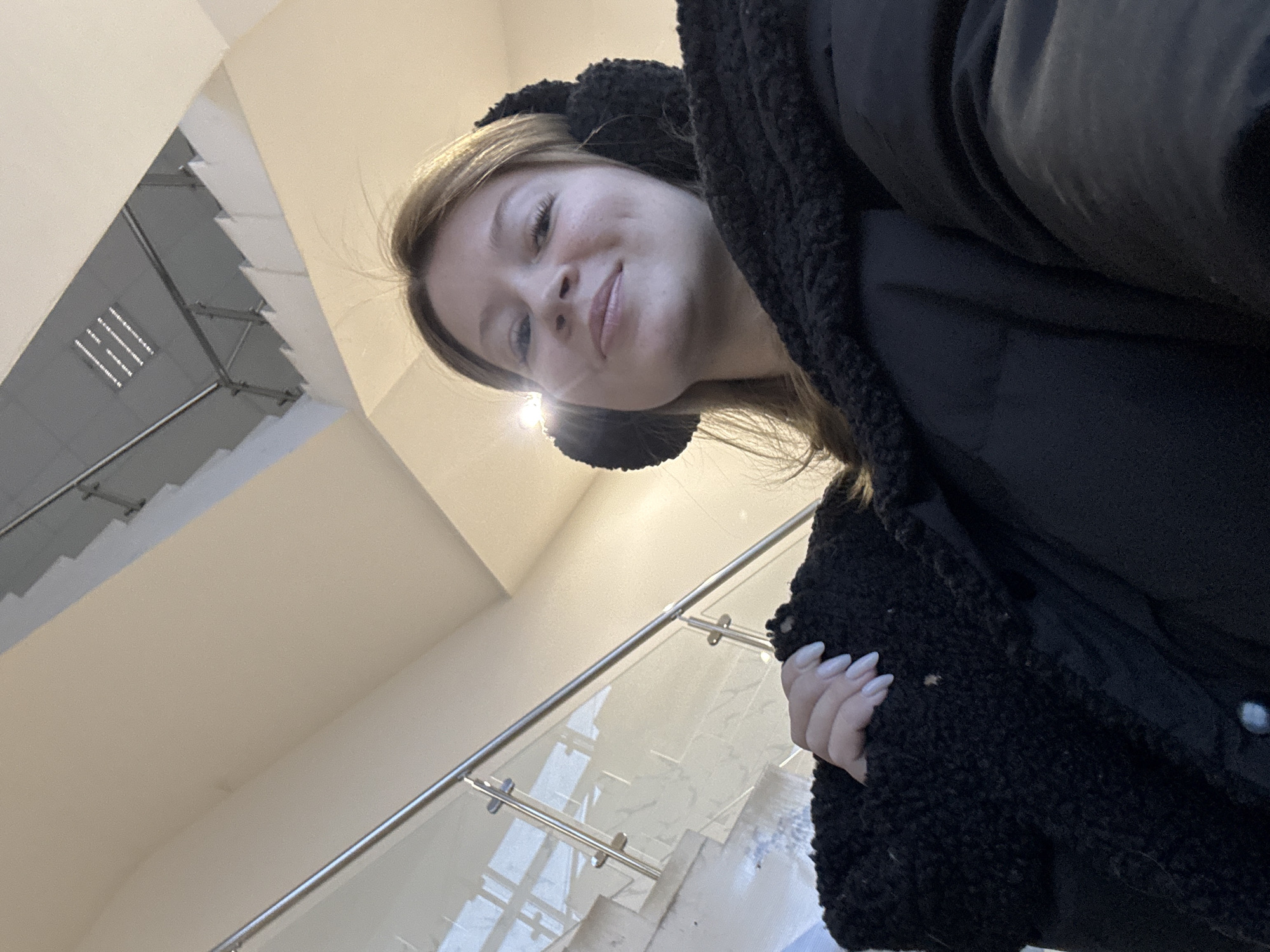 A woman wearing black earmuffs and a black coat stands inside a building with a staircase. The staircase features a glass railing with a metal support system. The location may be a public building in Chaikovsky, Russia, based on context. The woman is looking towards the camera. She has fair skin, light brown hair, and a slight smile. Her fingernails are painted white. The background includes a white ceiling with recessed lighting and the architecture of a staircase.