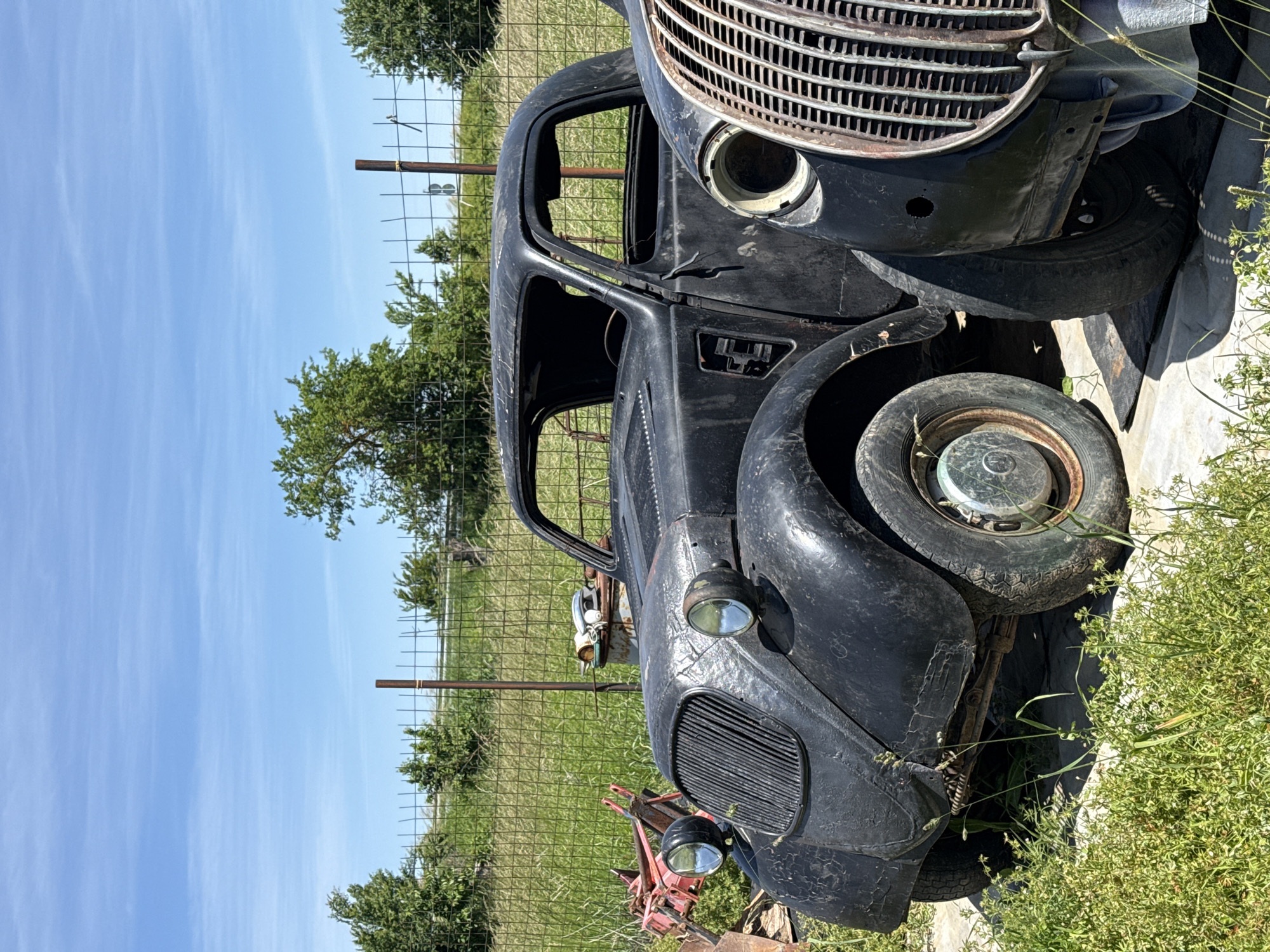 The image depicts a derelict, vintage black vehicle in a rural setting. The car appears to be in a state of disrepair, with visible rust and missing components. It is located near a wire fence, with a grassy field and trees in the background. The sky is blue, suggesting a clear day. The specific location is Omurtag, Bulgaria, based on the prompt.
