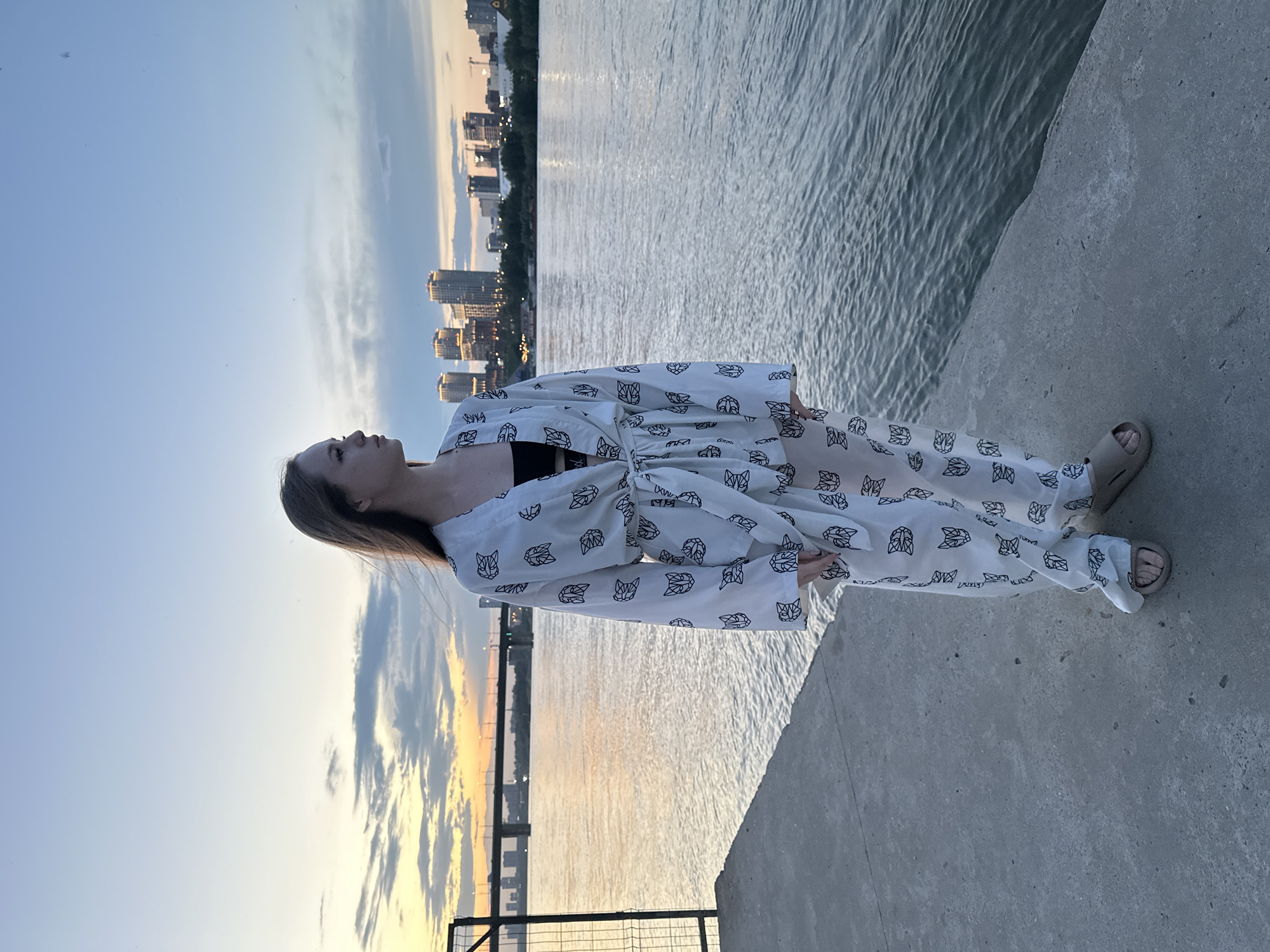 A young woman stands on a concrete pier overlooking a body of water, with a city skyline in the distance. She is wearing a white robe with a black cat-ear pattern, a black top, and beige slides. The sky is a soft blue with hints of orange and yellow from the setting sun. The water is calm, with gentle ripples reflecting the light. A metal fence is visible to the left of the woman. The city skyline features several tall buildings illuminated by lights.