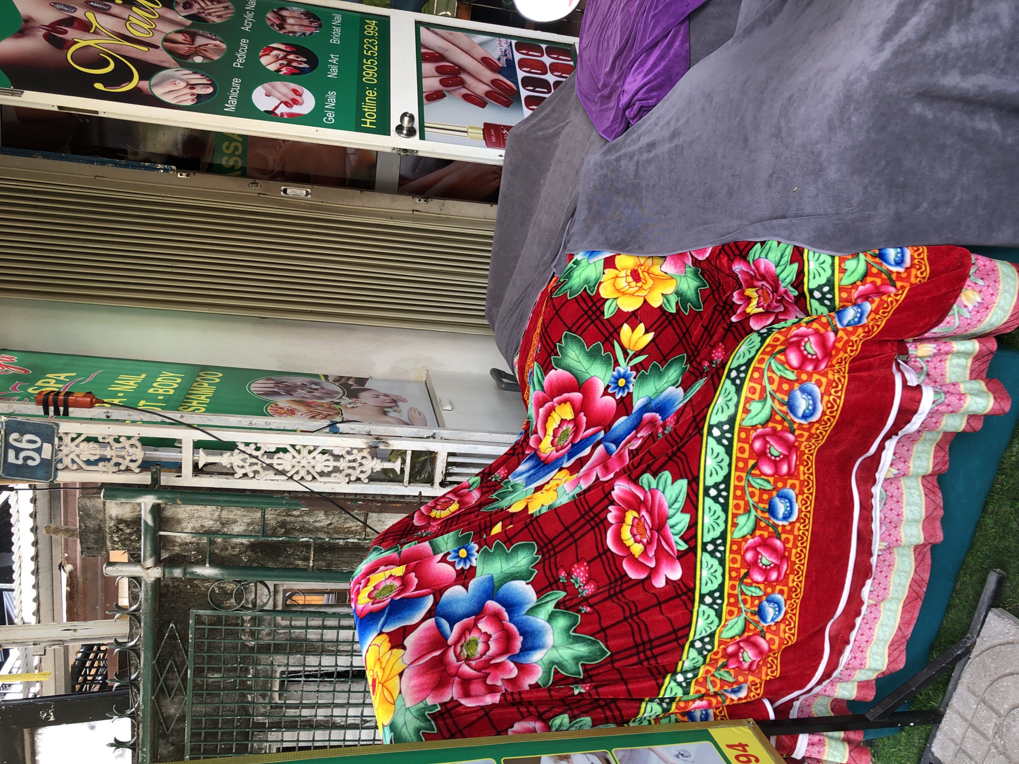 This is an outdoor, eye-level, medium shot of a massage bed covered with a red floral blanket and other fabric coverings, located in front of a spa in Da Nang, Vietnam. The spa appears to be on a street, with a closed metal rolling door to its right. A green sign to the left of the entrance lists services including "SPA," "NAIL," "BODY," and "SHAMPOO." To the right of the entrance is another sign with images of manicured hands and feet, and text for "Manicure," "Pedicure," "Acrylic Nail," "Gel Nails," "Nail Art," and "Bridal Nail." A phone number, "0905.523.994," is also visible on this sign. The massage bed itself is covered with dark grey fabric, with a purple fabric draped on top on the right. The red floral blanket is draped over the left side of the bed, featuring large pink, blue, and yellow flowers with green leaves, set against a red checkered background. The blanket has decorative borders with geometric patterns and smaller floral designs. In the background, a metal gate and a pillar with a house number "56" are visible. The overall impression is that the spa is closed or preparing for business.