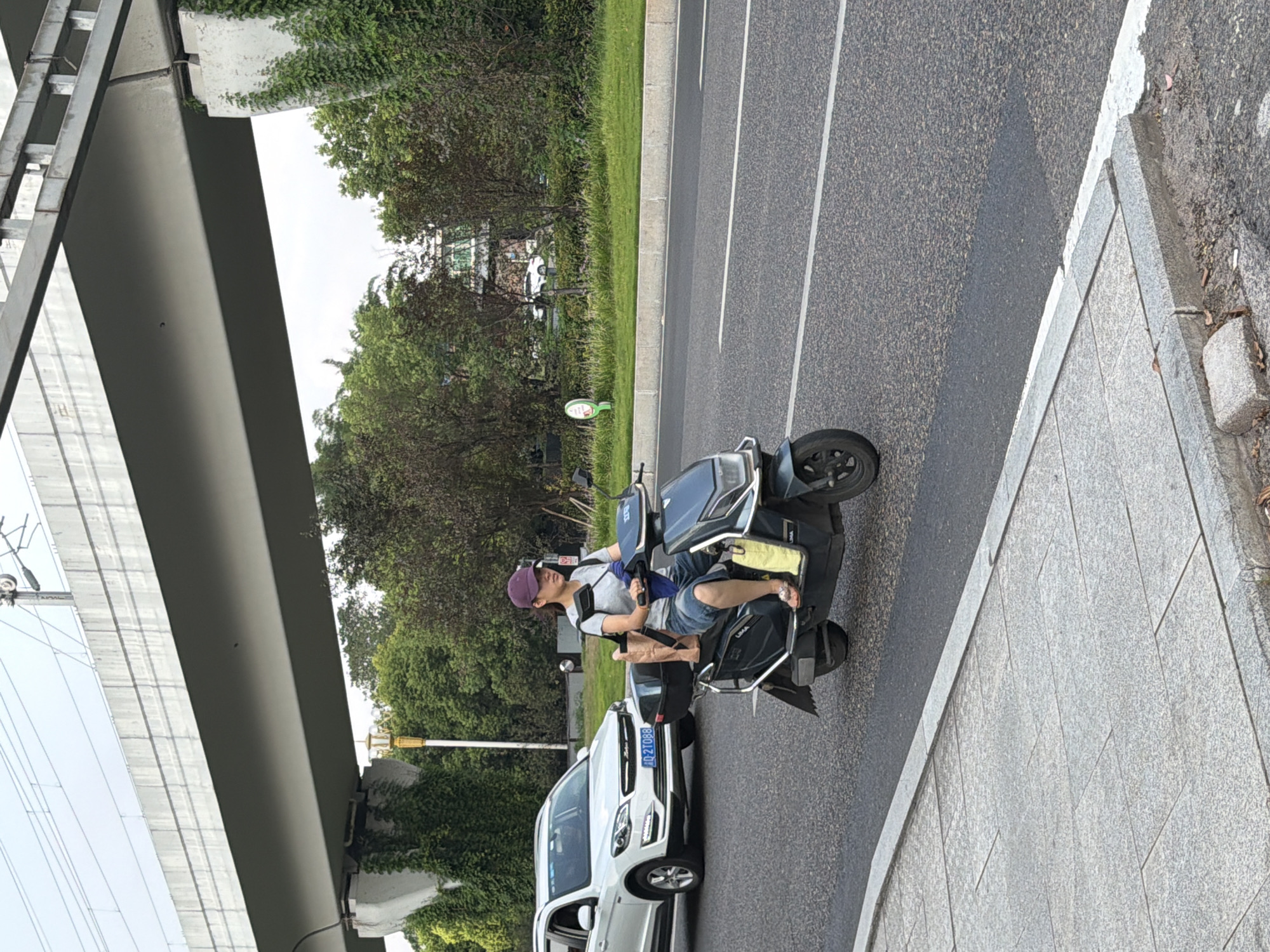A woman wearing a purple baseball cap, grey t-shirt, and denim shorts rides a dark blue electric scooter on a paved road. She is carrying a brown paper bag and a yellow bag. A white SUV with license plate "JD 2T088" is driving in the opposite direction on the left side of the road. An elevated concrete structure with electrical wires runs overhead. The scene is set in Jiangjin, China, with trees and a building visible in the background.
